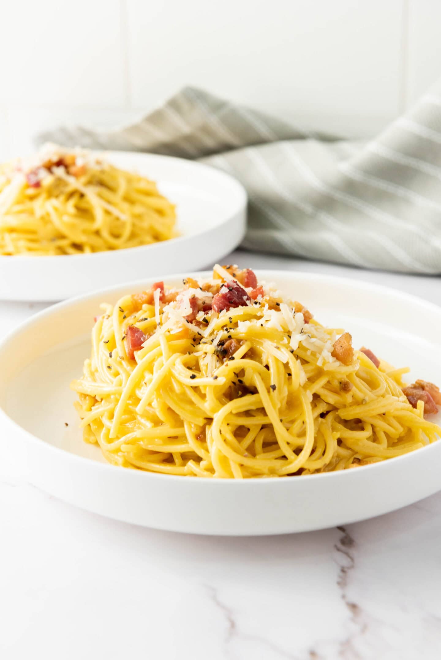 Two white plates with servings of authentic Spaghetti Carbonara, topped with grated cheese and bits of bacon, set on a white surface with a striped cloth in the background.