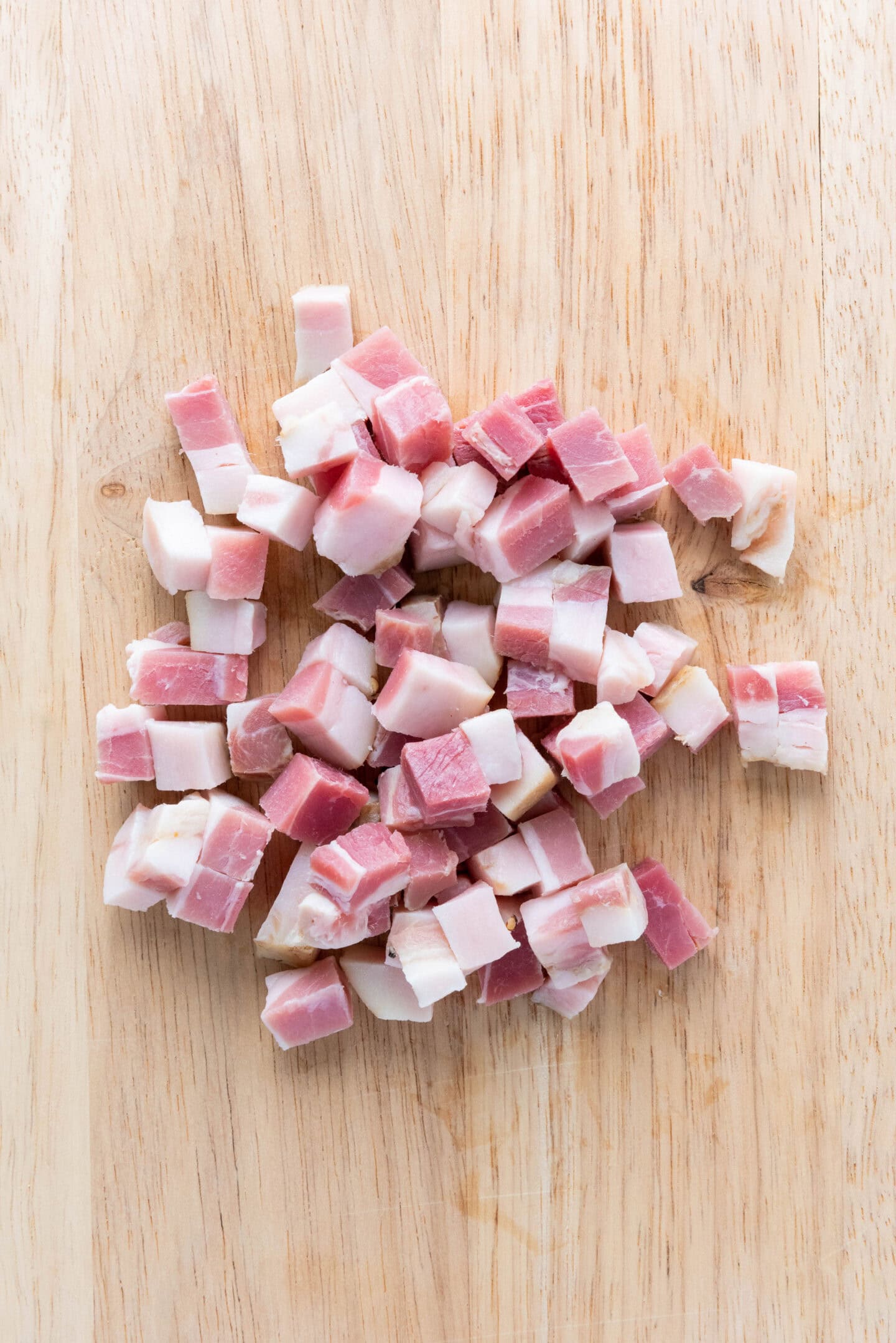 Small cubes of raw pancetta are scattered on a light wooden cutting board, ready to be the flavorful base for authentic Spaghetti Carbonara.