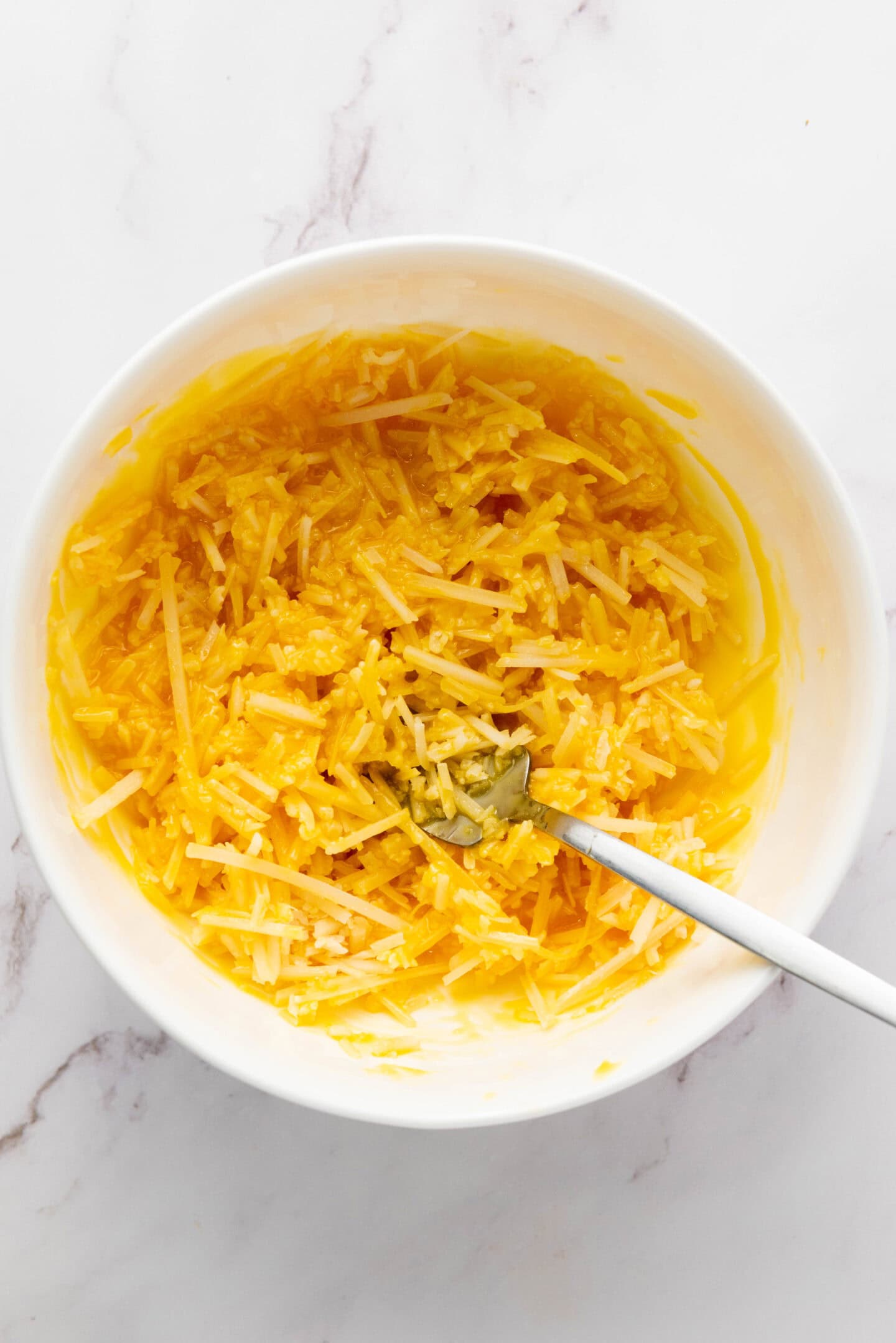A white bowl filled with a mixture of shredded yellow squash and cheese, reminiscent of an authentic Carbonara, with a metal spoon resting inside, on a white marble surface.