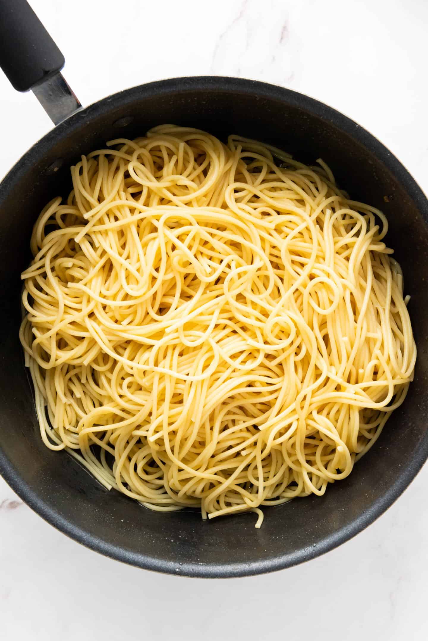 A black pot filled with cooked, plain spaghetti noodles on a white marble surface, ready to be transformed into an authentic Spaghetti Carbonara.