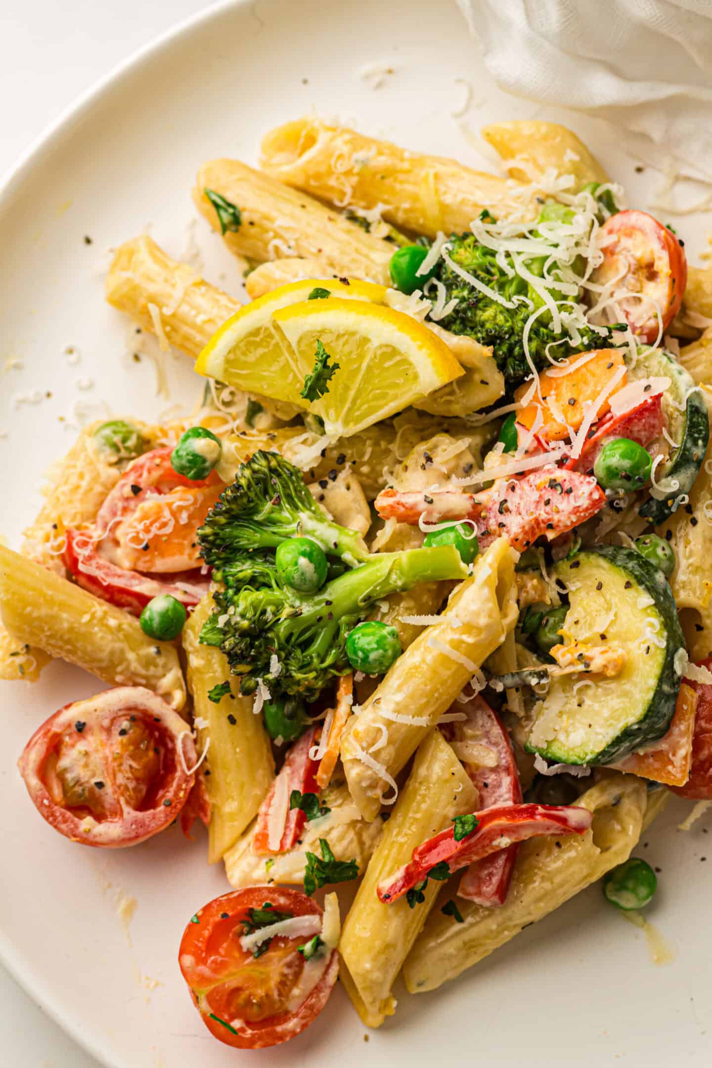 Plate of Pasta Primavera featuring penne tossed with assorted vegetables—broccoli, tomatoes, zucchini, and peas—garnished with grated cheese, lemon wedges, and fresh herbs for a vibrant vegetable pasta dish.