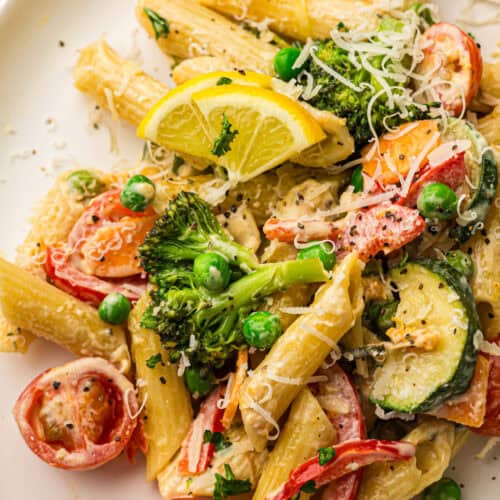 Plate of Pasta Primavera featuring penne tossed with assorted vegetables—broccoli, tomatoes, zucchini, and peas—garnished with grated cheese, lemon wedges, and fresh herbs for a vibrant vegetable pasta dish.