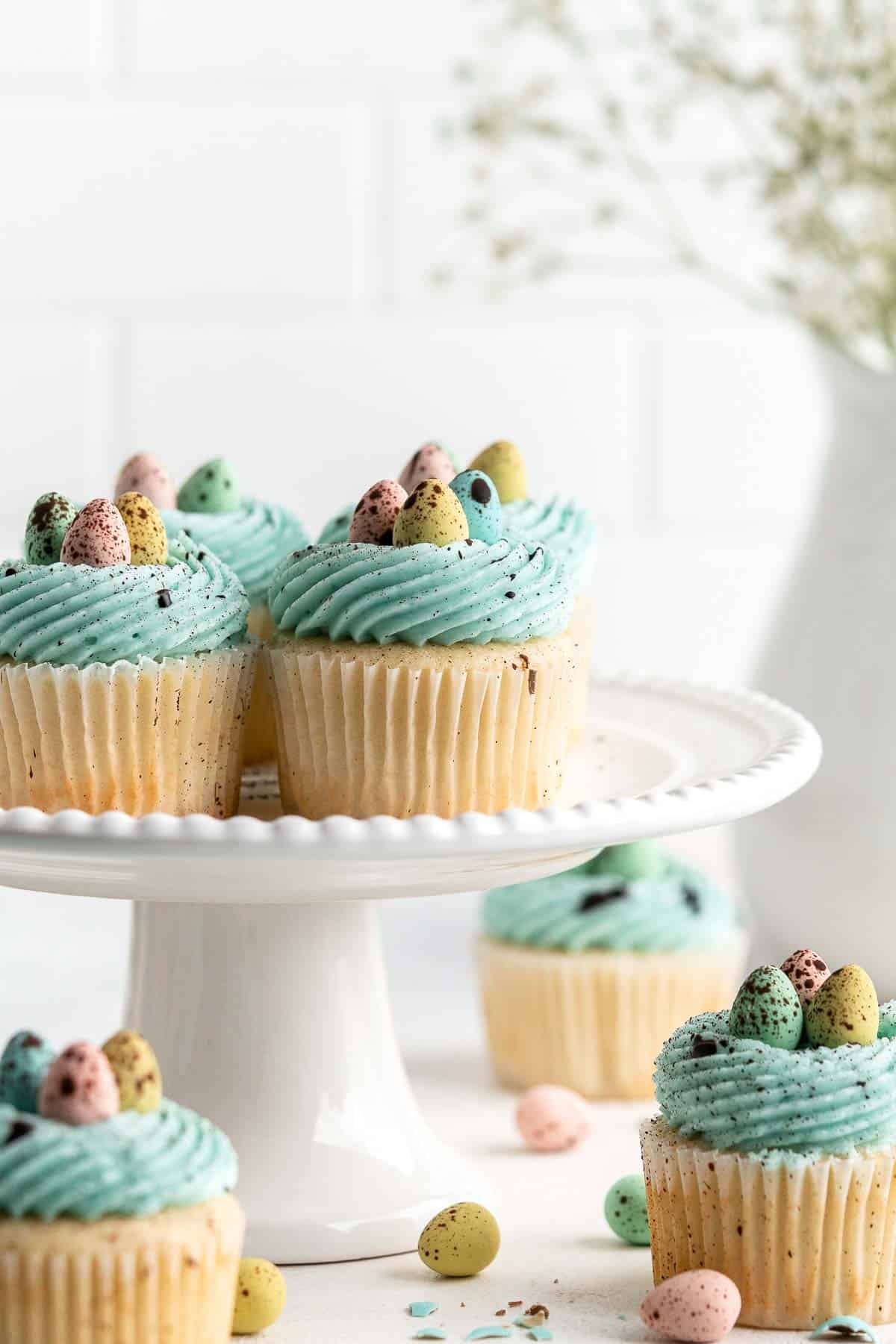 Cupcakes with blue frosting and candy eggs on top are arranged on a white cake stand, making them perfect Easter Party Food, with more cupcakes and festive Easter Snacks in the background.