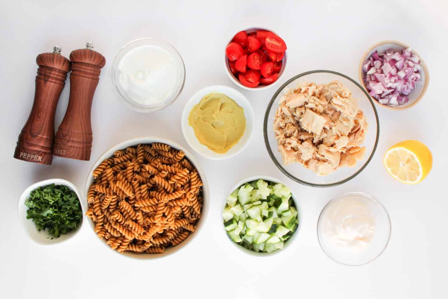 Ingredients for a healthy Tuna Pasta Salad arranged on a white surface, featuring fusilli pasta, tuna, cucumber, cherry tomatoes, red onion, herbs, lemon, mustard, Greek yogurt, sauces, salt, and pepper.