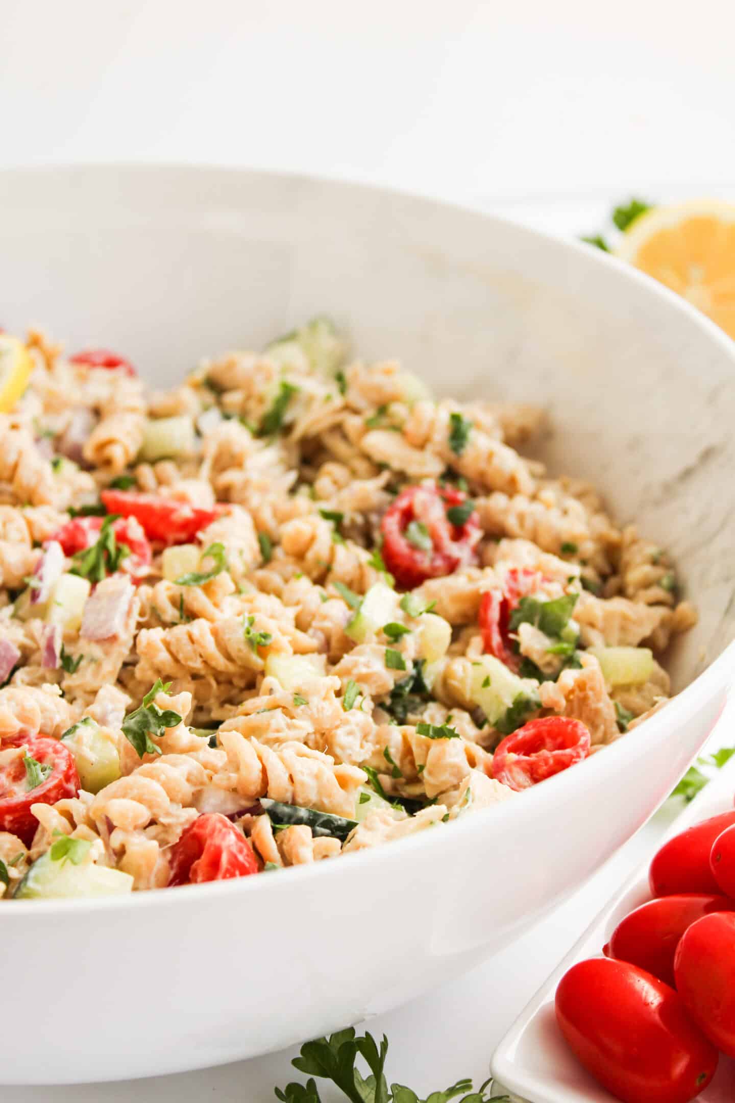A large white bowl filled with healthy pasta salad containing rotini, cherry tomatoes, cucumber, herbs, and a creamy dressing; fresh tomatoes are placed beside the bowl.