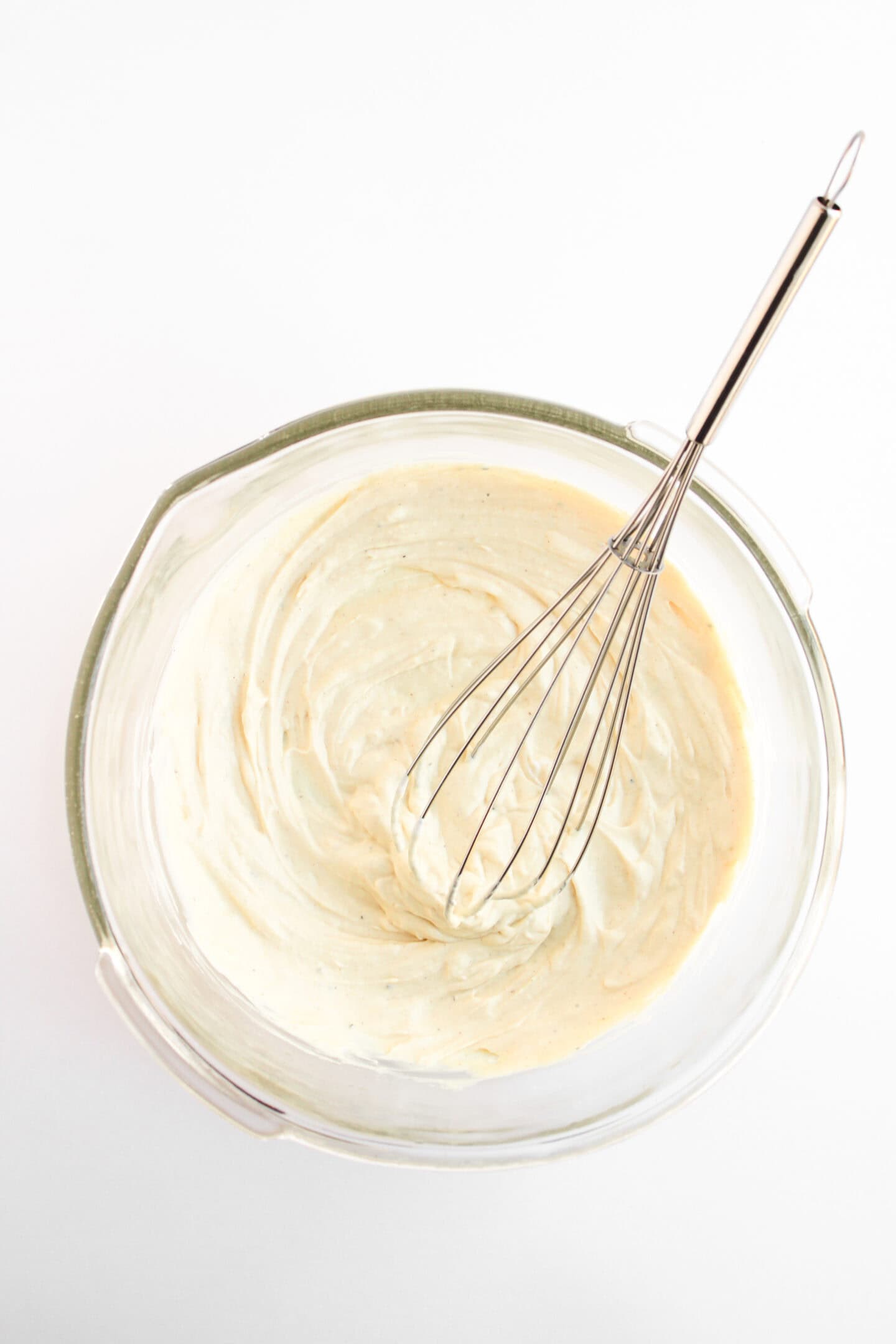 A glass mixing bowl filled with creamy, healthy batter—made with Greek yogurt—and a metal whisk resting inside, set against a white background.