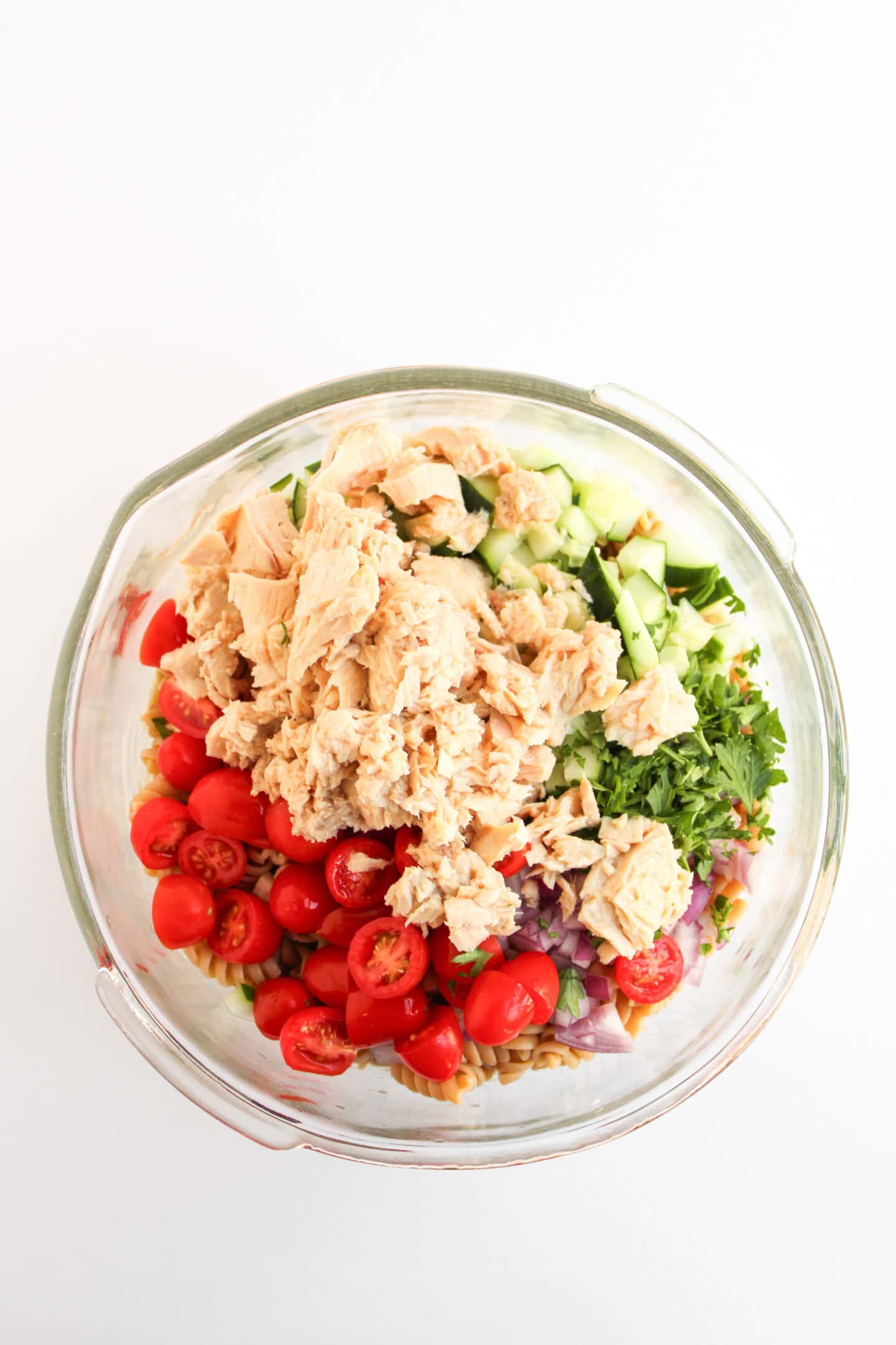 A glass bowl filled with chopped cucumbers, cherry tomatoes, red onions, parsley, and chunks of tuna creates a healthy dish on a white background.