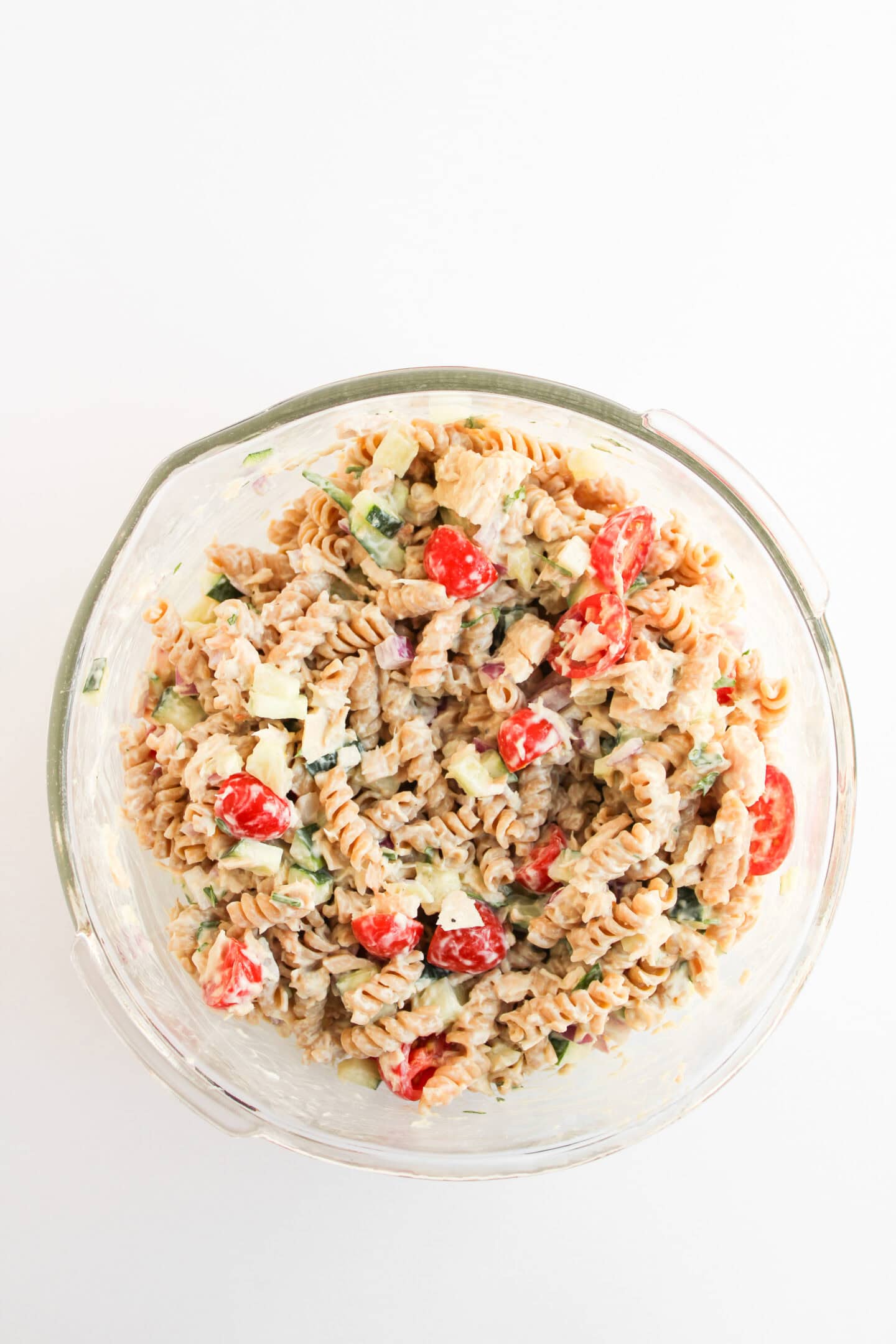 A glass bowl filled with healthy rotini pasta salad featuring cherry tomatoes, diced cucumbers, and other mixed vegetables in a creamy Greek yogurt dressing, viewed from above.