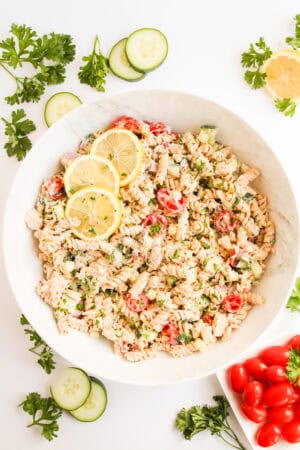 A bowl of healthy tuna pasta salad with cherry tomatoes, cucumbers, herbs, and lemon slices, surrounded by fresh parsley, cucumber slices, grape tomatoes, and a creamy Greek yogurt dressing.