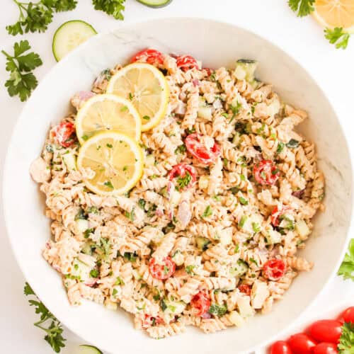 A bowl of healthy tuna pasta salad with cherry tomatoes, cucumbers, herbs, and lemon slices, surrounded by fresh parsley, cucumber slices, grape tomatoes, and a creamy Greek yogurt dressing.