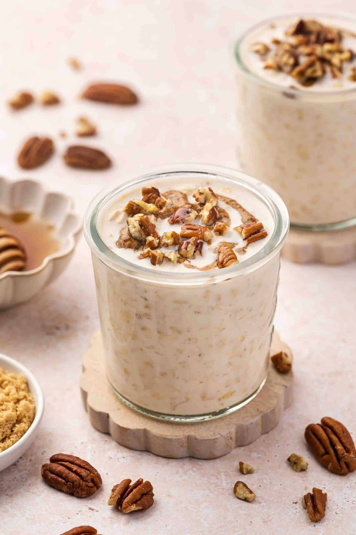 A glass of creamy oatmeal, perfect for a healthy breakfast, topped with chopped pecans on a coaster, with another similar glass—oats made without chia seeds—honey, and brown sugar in the background.