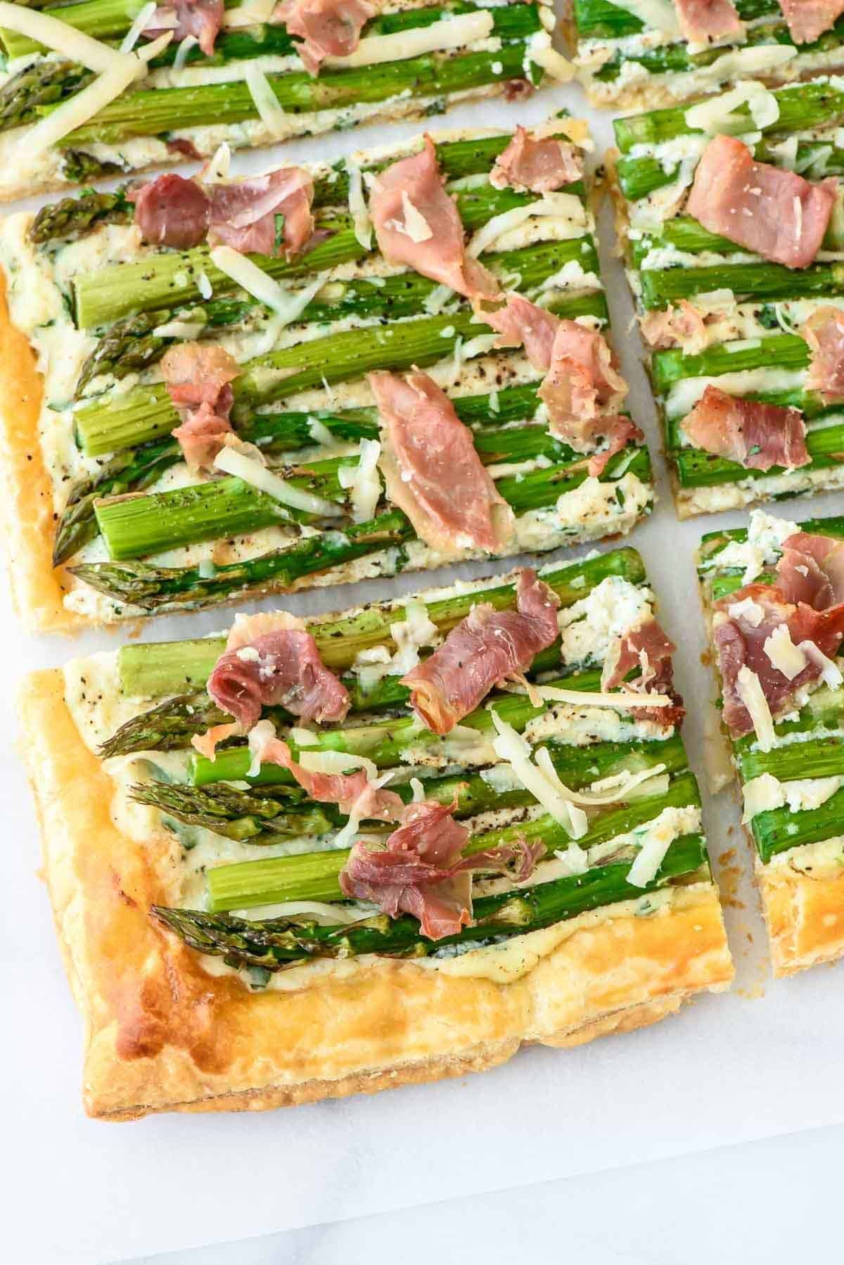 Rectangular pieces of puff pastry topped with asparagus, prosciutto, and shredded cheese are arranged closely together—perfect as elegant spring appetizers.