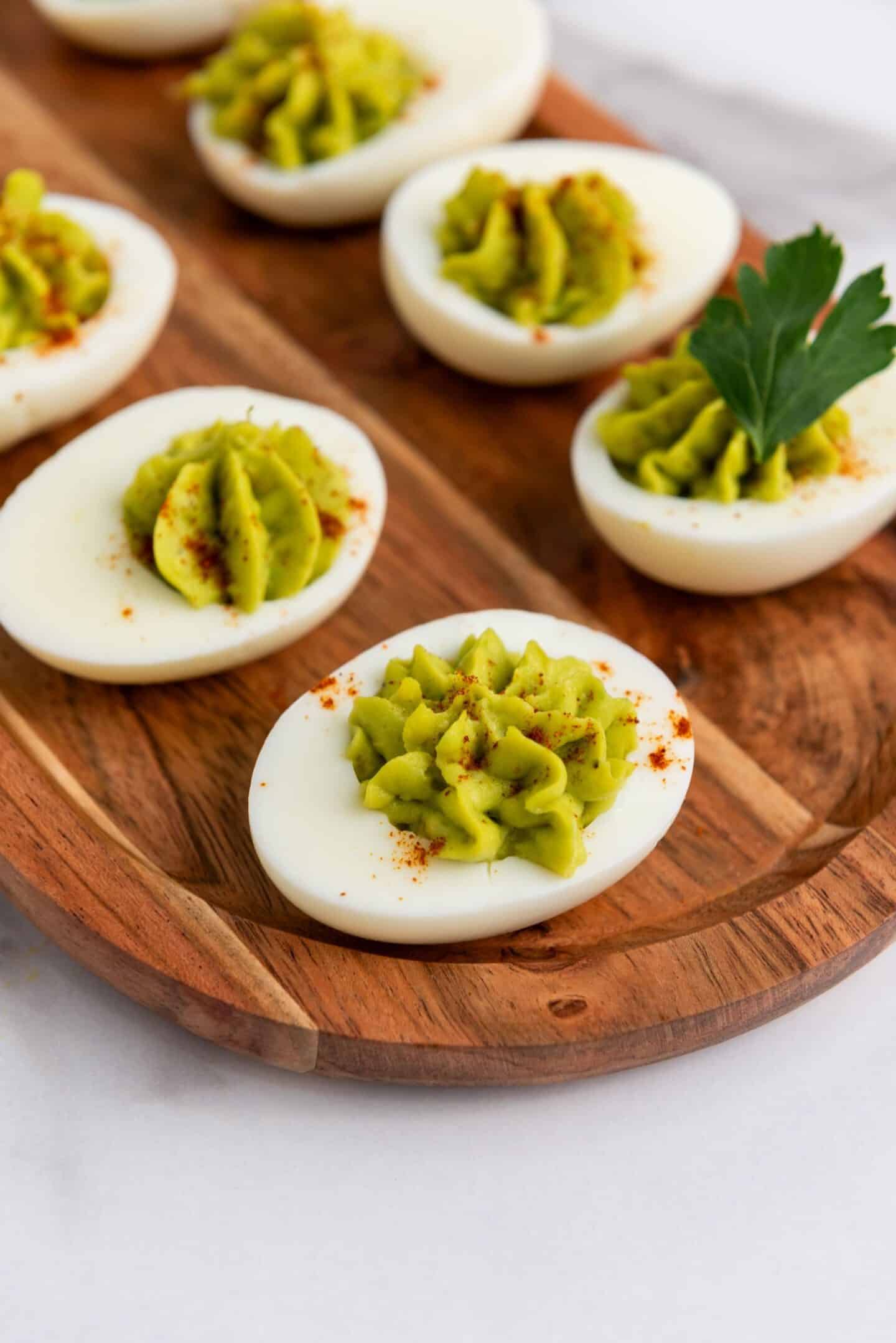 Deviled eggs on a wooden tray, filled with green avocado mixture and sprinkled with paprika; one is garnished with a parsley leaf—perfect as vibrant Spring Appetizers.