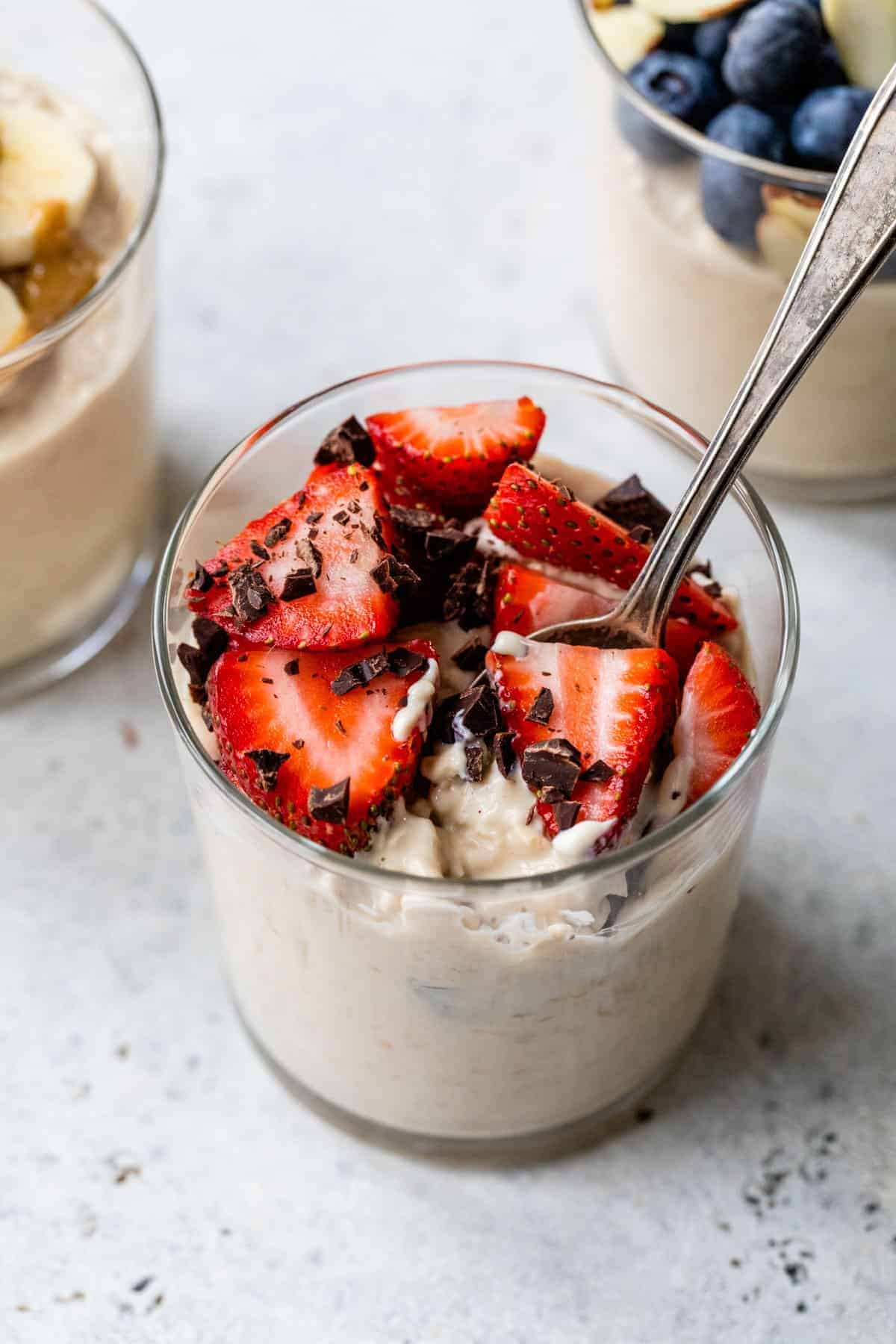 A glass of oats breakfast without chia seeds, topped with sliced strawberries and chocolate pieces, with a spoon inside. Two similar glasses featuring different overnight oats recipes are in the background.