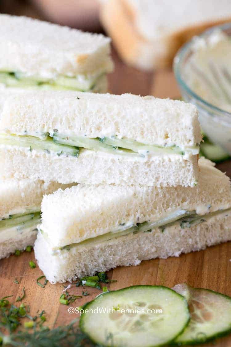 Three rectangular cucumber tea sandwiches made with white bread, cream cheese, and cucumber slices are stacked on a wooden surface with scattered herbs—perfect spring appetizers for any gathering.