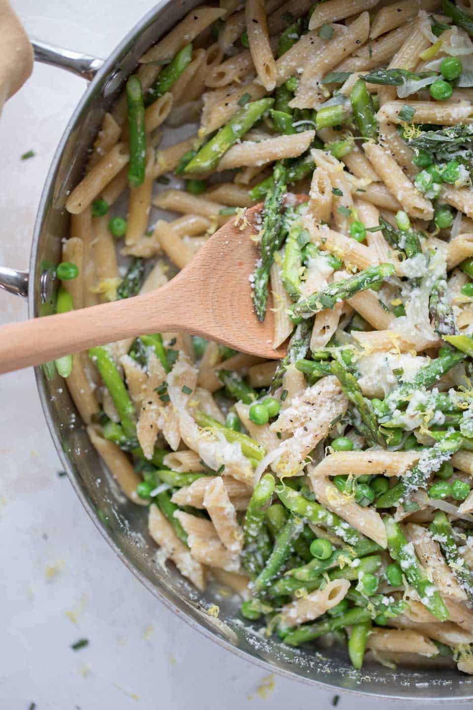A skillet filled with penne pasta, asparagus, peas, and a creamy white sauce, being stirred with a wooden spoon—a delicious addition to your spring pasta recipes.