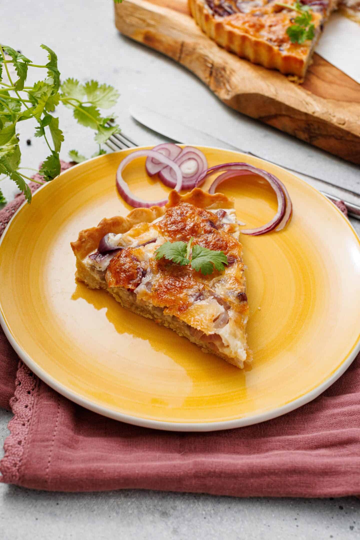 A slice of savory tart with vegetables and herbs, perfect for spring appetizers, served on a yellow plate and garnished with red onion rings and cilantro.