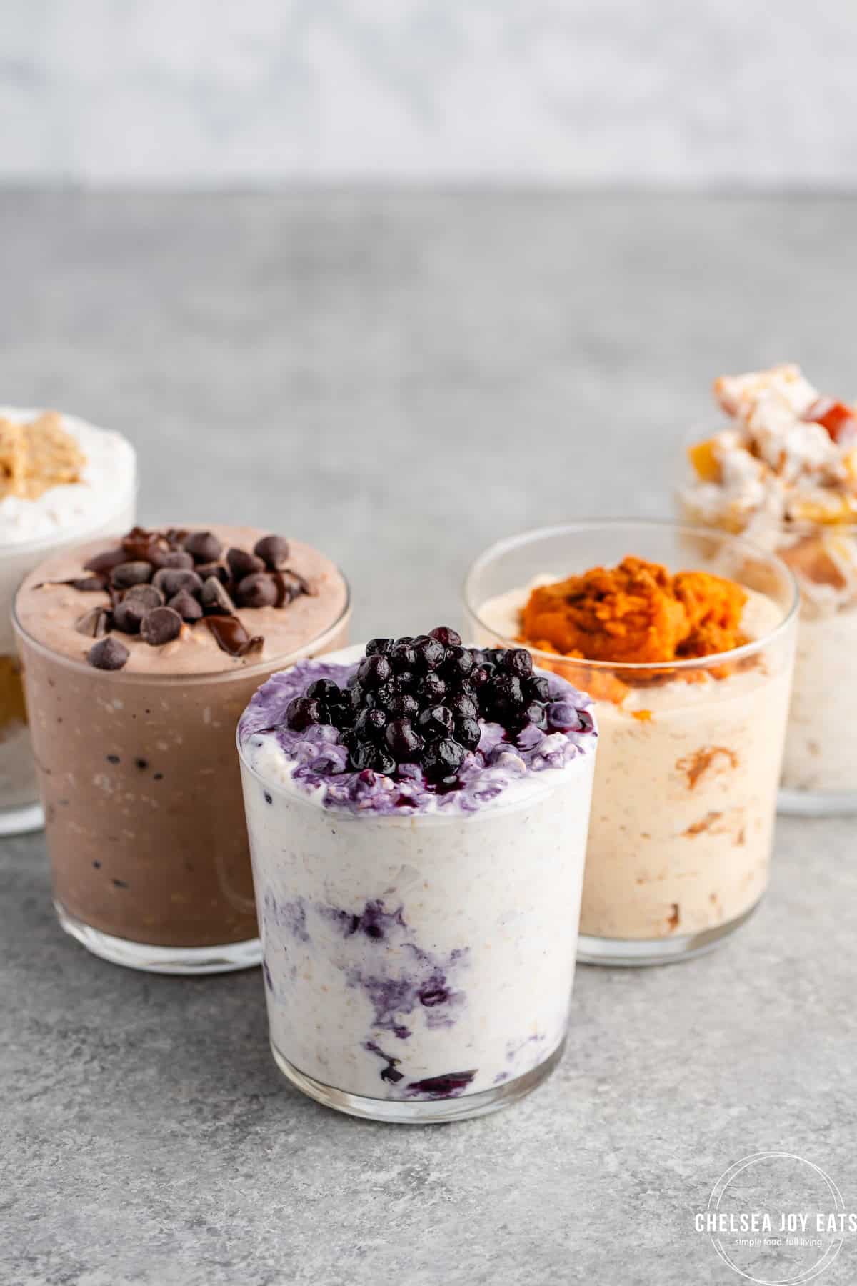Five glass cups filled with different chia-free overnight oats are arranged on a gray surface, each topped with ingredients like blueberries, chocolate chips, and pumpkin puree—delicious overnight oats recipes without chia seeds.