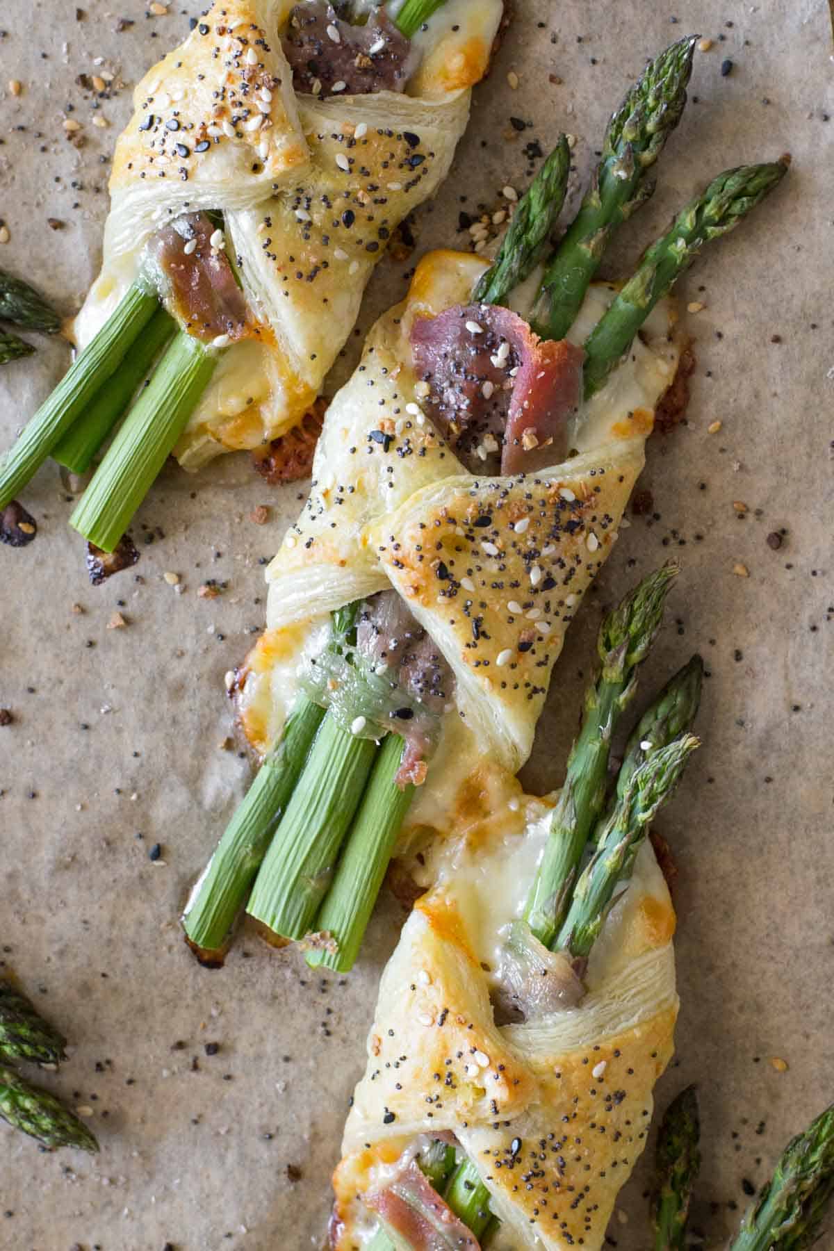 Three puff pastry bundles filled with asparagus, cheese, and prosciutto, topped with seasoning, make perfect Spring Appetizers when arranged on parchment paper.