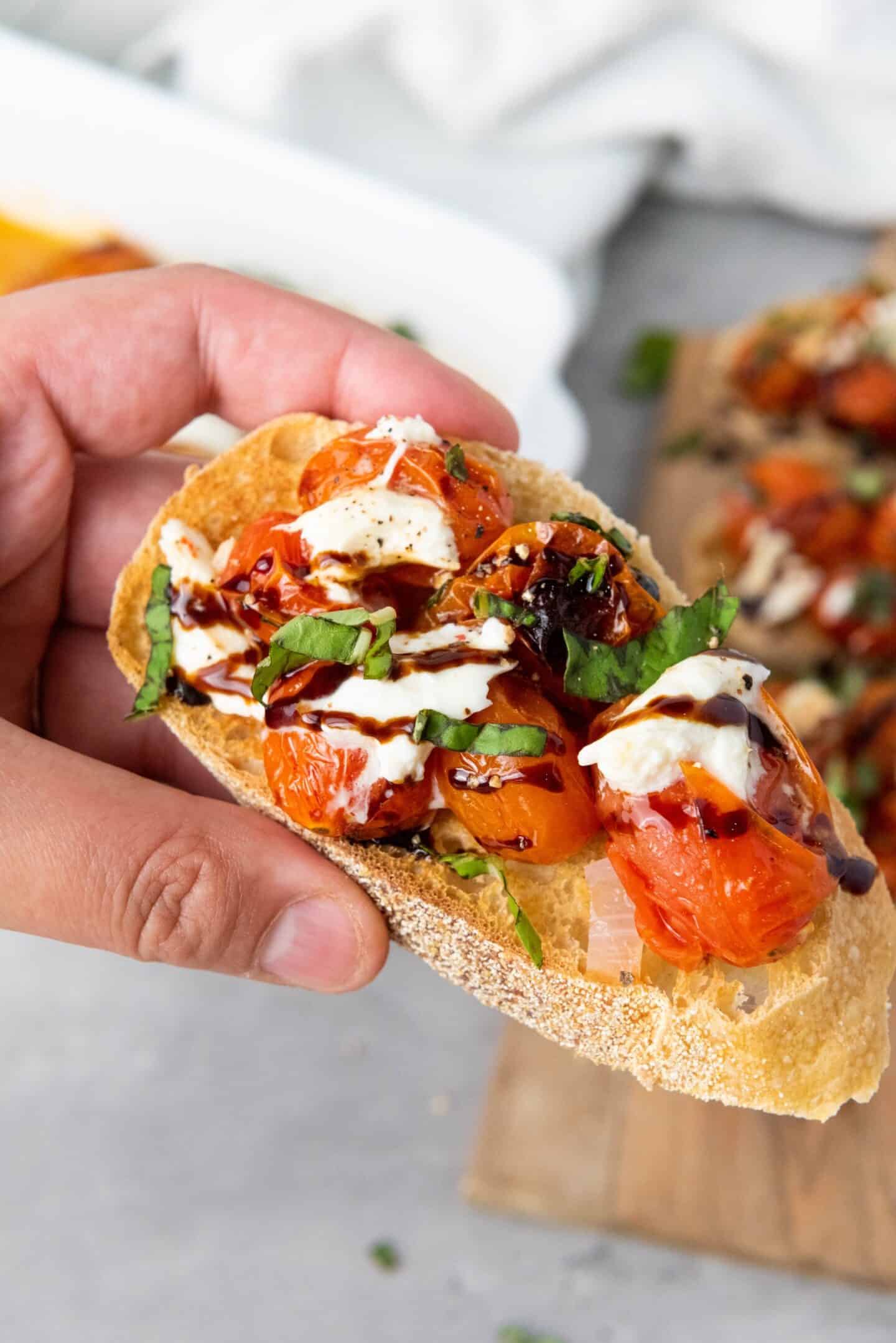 A hand holds a slice of toasted bread topped with roasted cherry tomatoes, fresh basil, mozzarella, and a drizzle of balsamic glaze—perfect for serving as spring appetizers.