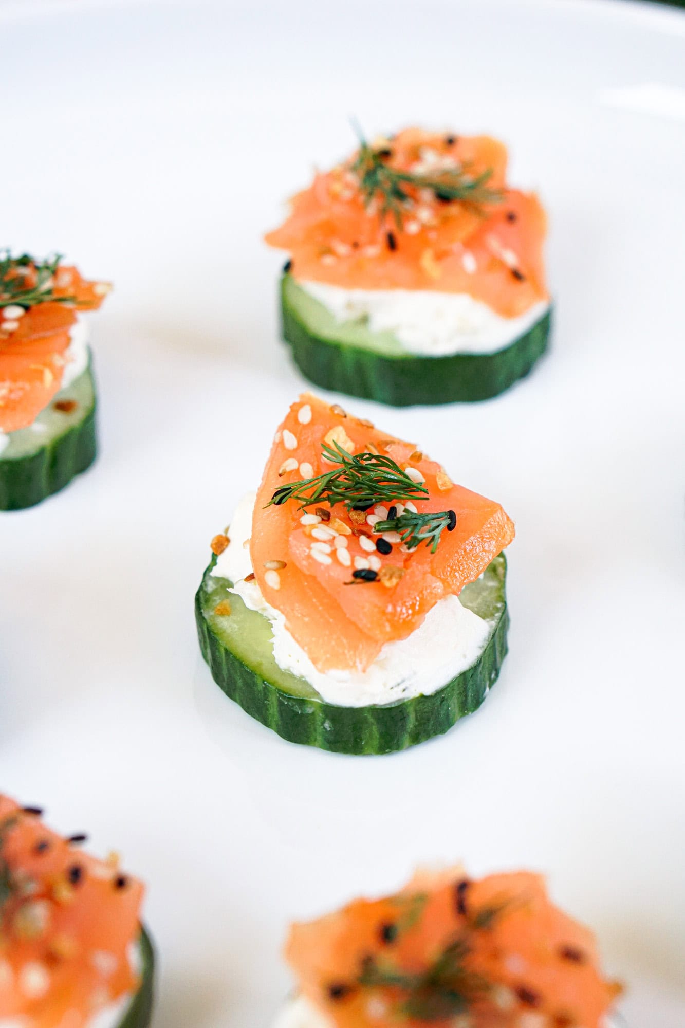 Slices of cucumber topped with cream cheese, smoked salmon, dill, and sesame seeds make fresh spring appetizers, beautifully arranged on a white plate.
