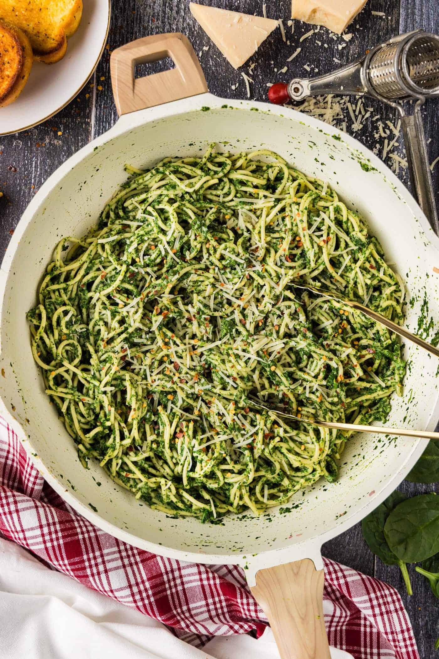 A skillet filled with spaghetti coated in green pesto sauce, topped with grated cheese, showcases one of the freshest spring pasta recipes, with utensils resting in the pasta and kitchen items in the background.