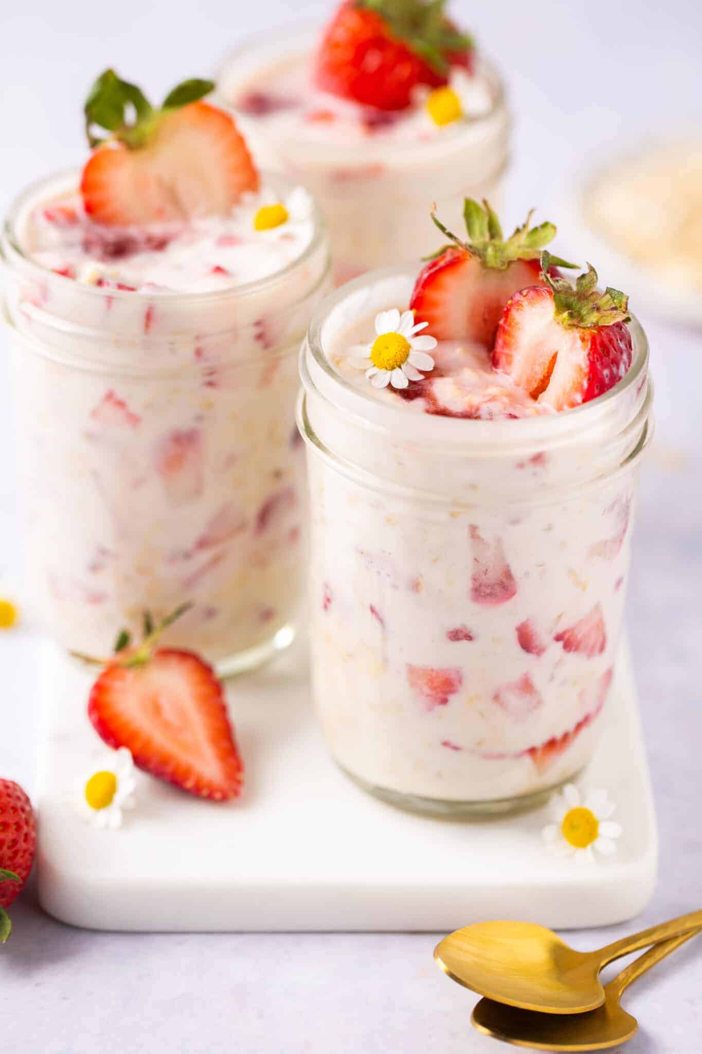 Three glass jars filled with strawberry yogurt parfaits, inspired by chia-free overnight oats, topped with fresh strawberry halves and small white flowers, placed on a white board next to gold spoons.
