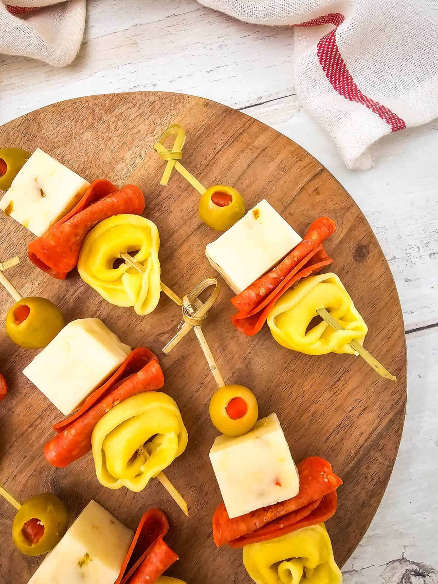 Wooden board with skewers featuring cheese cubes, slices of pepperoni, tortellini pasta, and green olives, arranged neatly for serving—perfect for spring appetizers.