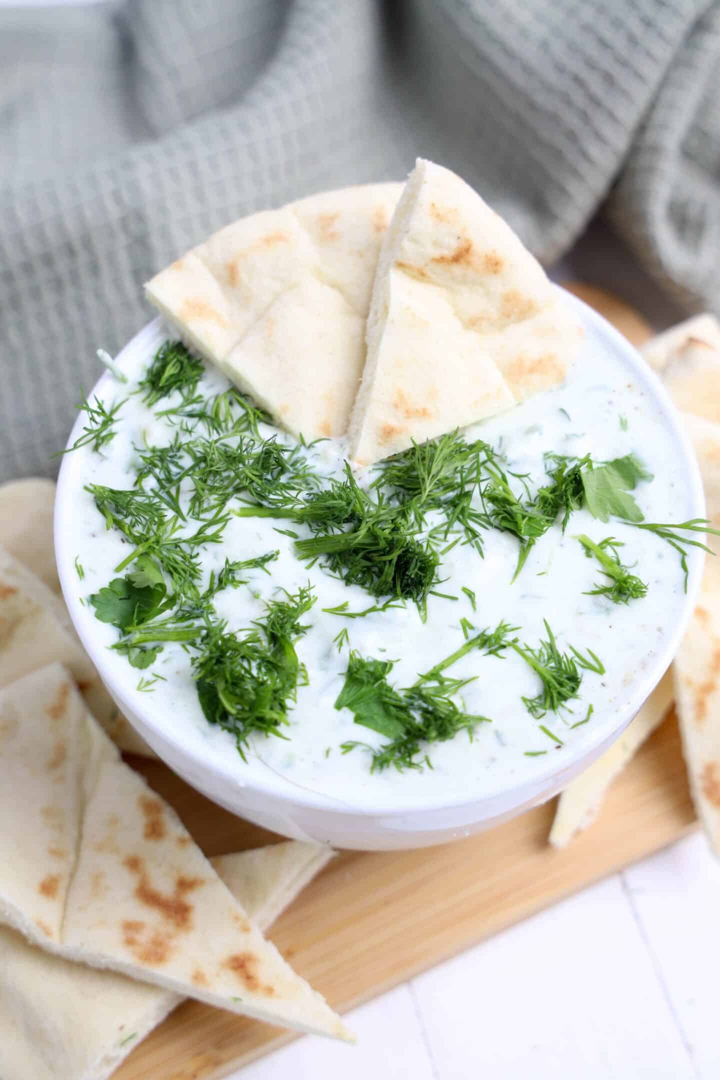 A bowl of tzatziki sauce garnished with fresh herbs, served with pieces of pita bread on a wooden board—perfect for light spring appetizers.