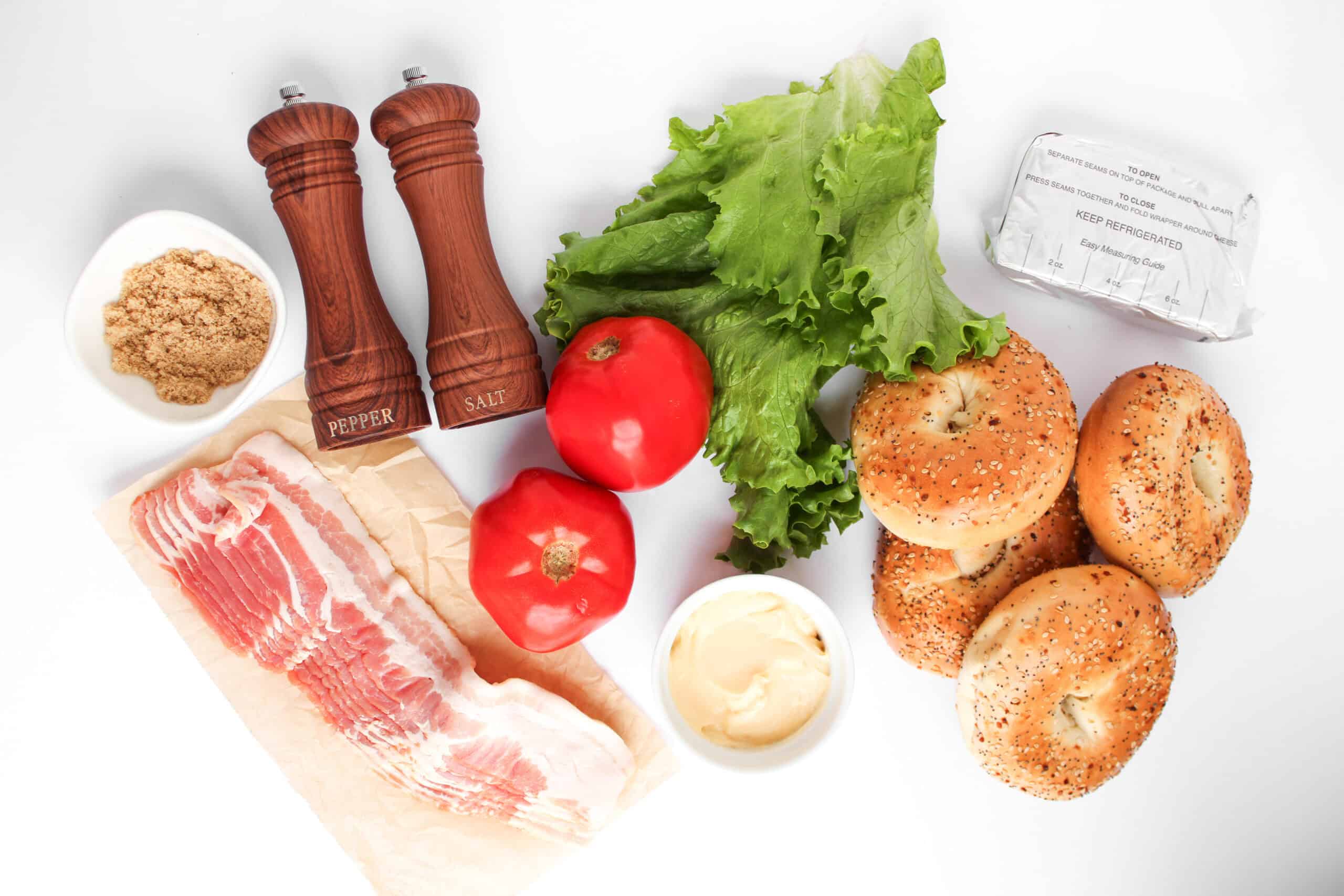 BLT Bagels are made with bagels, lettuce, tomatoes, bacon, cream cheese, mayonnaise, brown sugar, salt, and pepper arranged on a white surface.