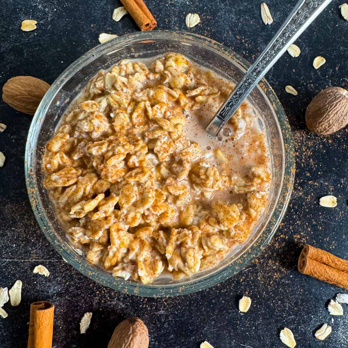 A bowl of oatmeal with milk and cinnamon, surrounded by whole nutmeg, cinnamon sticks, and scattered oats on a dark surface. Perfect for those seeking easy breakfast ideas or overnight oats recipes without chia seeds. A spoon is in the bowl.