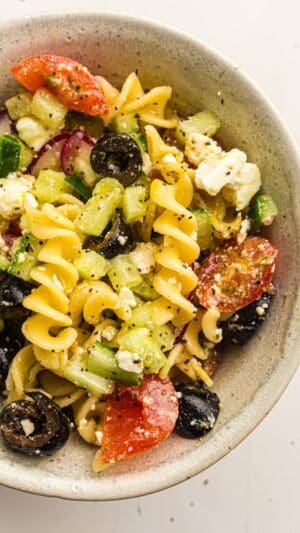A bowl of Greek Pasta Salad with rotini, cherry tomatoes, cucumbers, black olives, red onions, and crumbled feta cheese, all seasoned with herbs and black pepper.