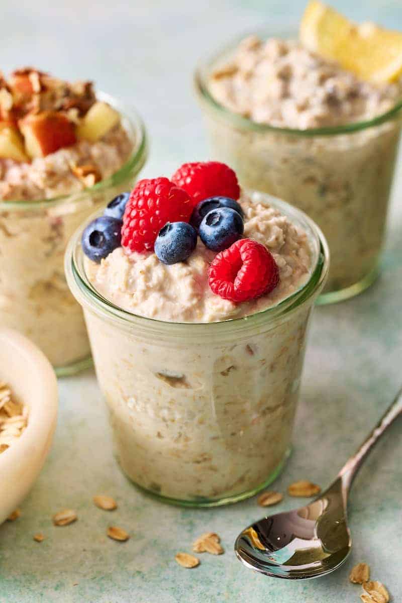 Glass jars filled with overnight oats recipes, without chia seeds, are topped with fresh berries and arranged on a table alongside a spoon and oat flakes.