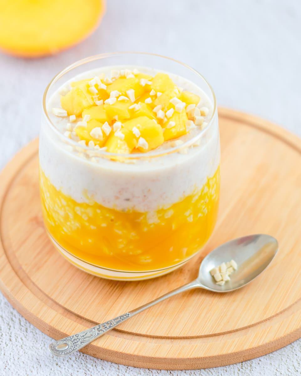 A glass of mango chia pudding topped with diced mango and chopped nuts sits on a round wooden tray beside a spoon—a perfect healthy breakfast option.