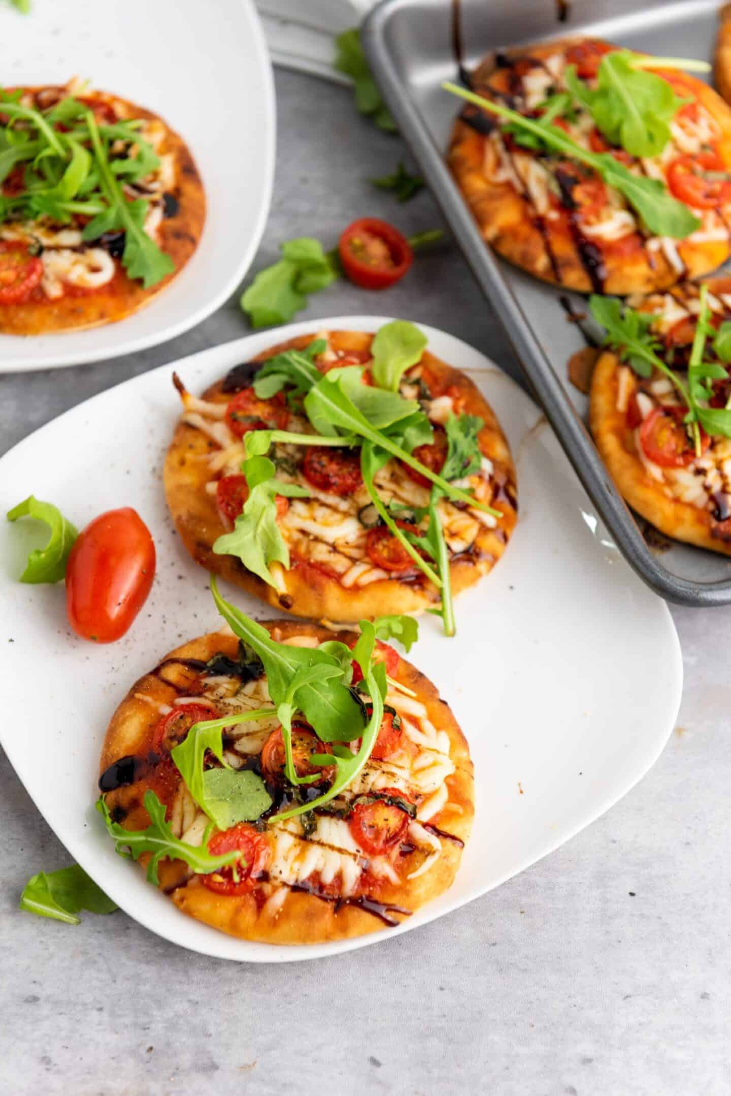 These small personal pizzas, perfect as spring appetizers, are topped with arugula, cherry tomatoes, cheese, and balsamic glaze, served on a white plate and baking tray on a light gray surface.