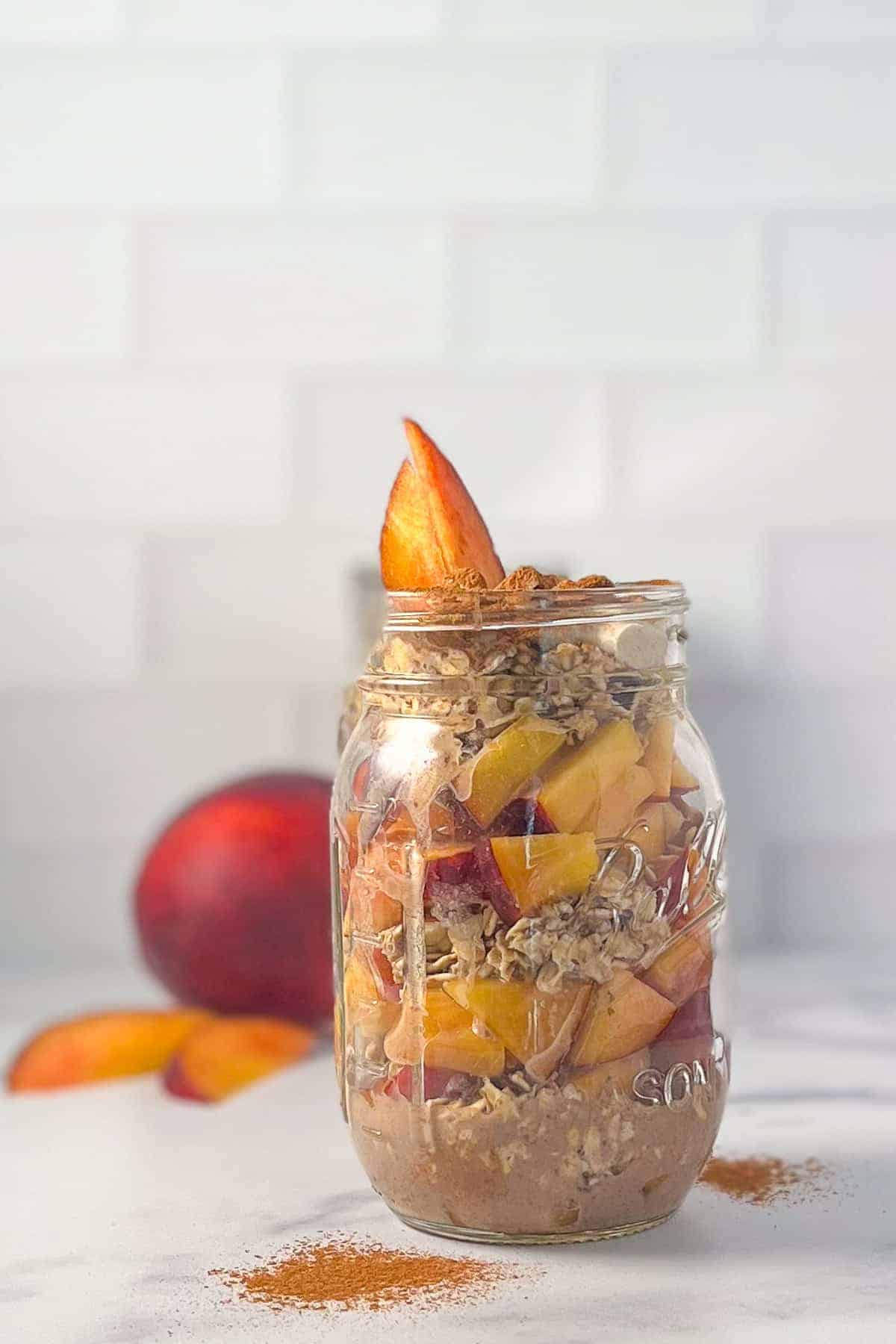 A mason jar filled with layers of sliced peaches, oats, and a brown mixture—without chia seeds—topped with peach slices and sprinkled with cinnamon. A delicious idea for those seeking new overnight oats recipes.
