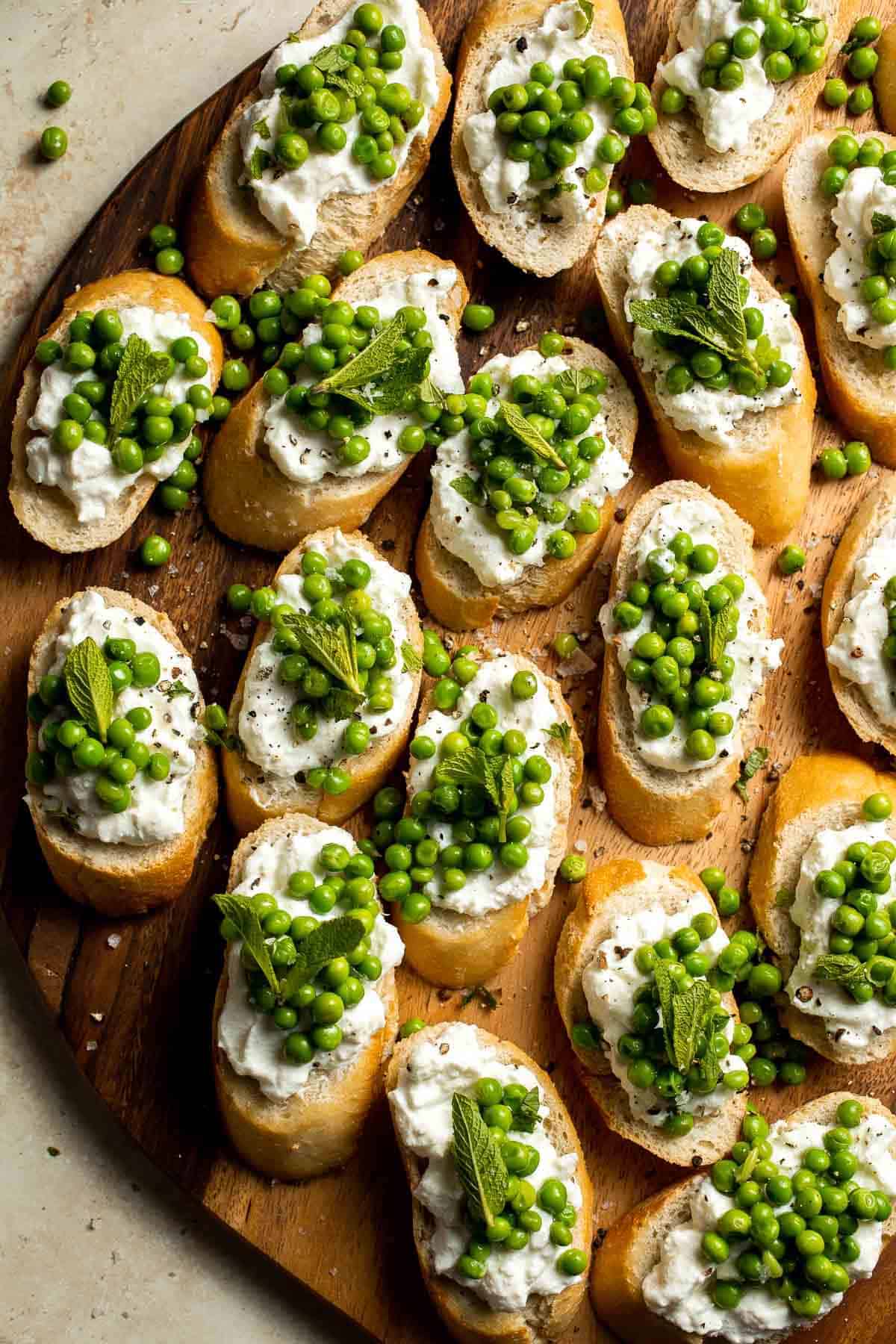 Crostini topped with ricotta cheese, green peas, and fresh mint leaves arranged on a wooden serving board—perfect as light and refreshing spring appetizers.