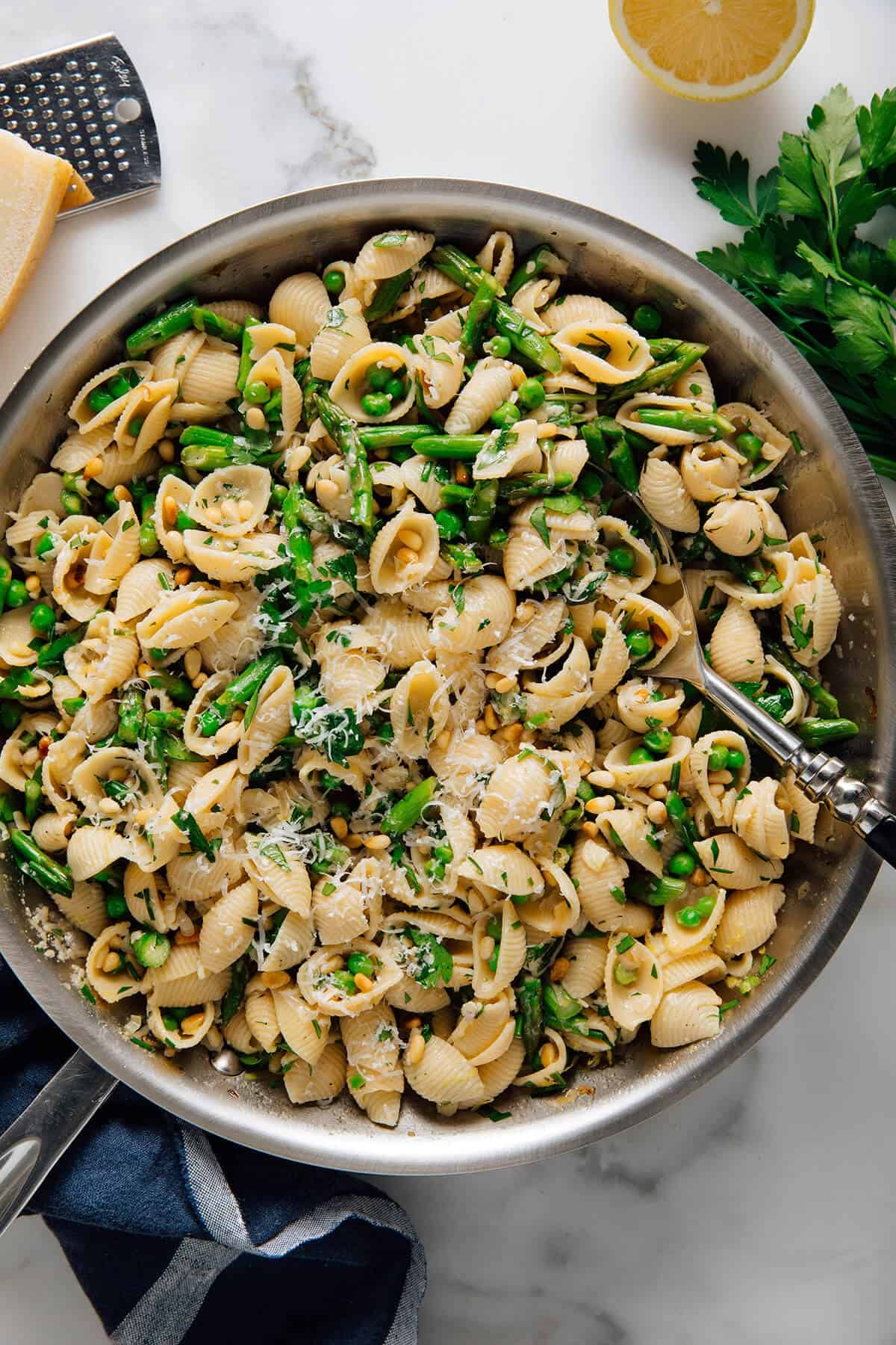 A skillet with shell pasta, green beans, peas, pine nuts, parsley, and grated cheese—perfect for spring pasta recipes. A serving spoon rests nearby, with lemon, parsley, and cheese on the side.