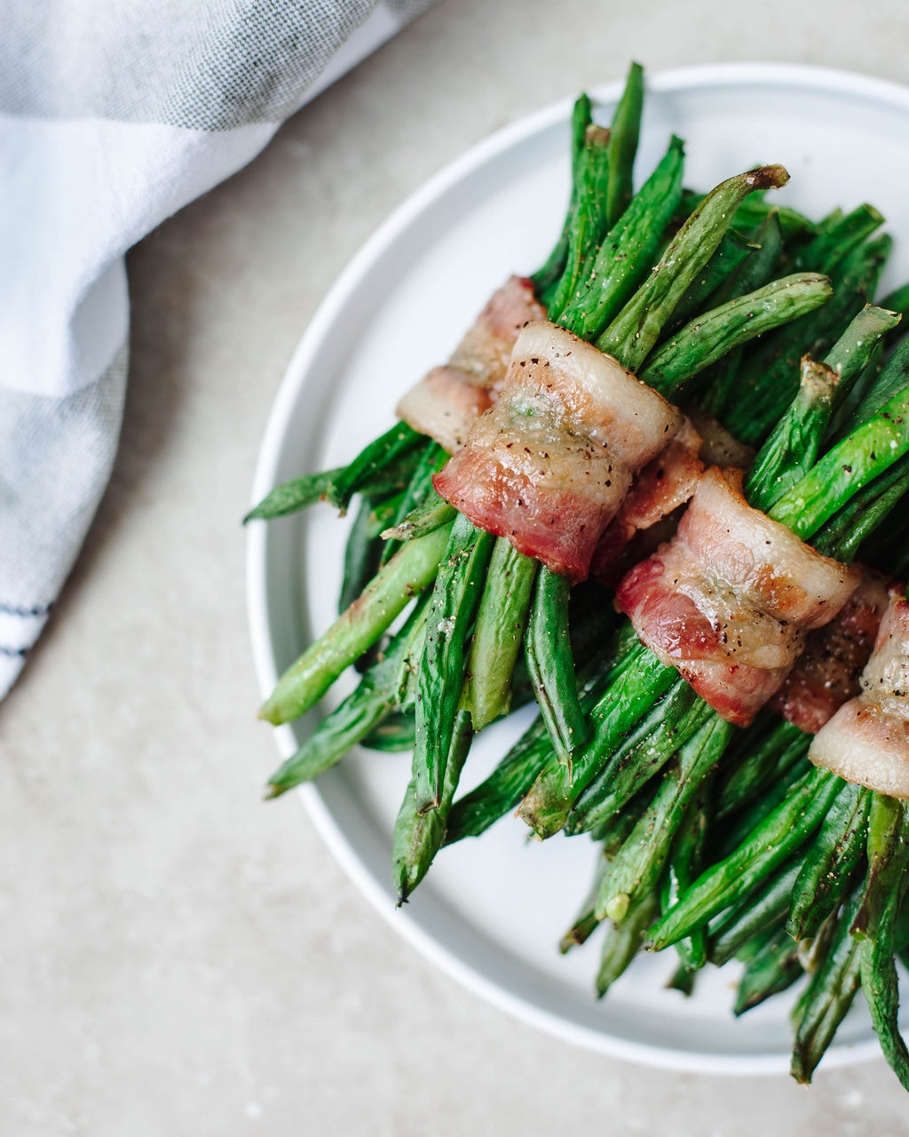 A white plate with bundles of green beans wrapped in bacon, placed on a light surface with part of a white cloth visible in the corner—perfect for serving as one of your summer vegetable side dishes.