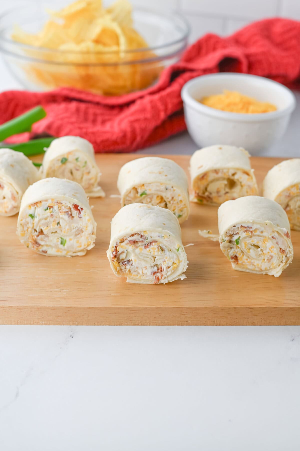 Several pinwheel tortilla roll-ups filled with a creamy mixture are arranged on a wooden board—perfect for Sports Dinners. In the background, there is a bowl of chips and a small bowl of shredded cheese.