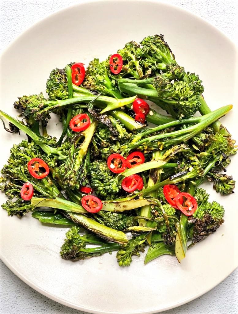 A plate of grilled broccolini garnished with sliced red chili peppers on a white surface—an ideal addition to your summer vegetable side dishes.