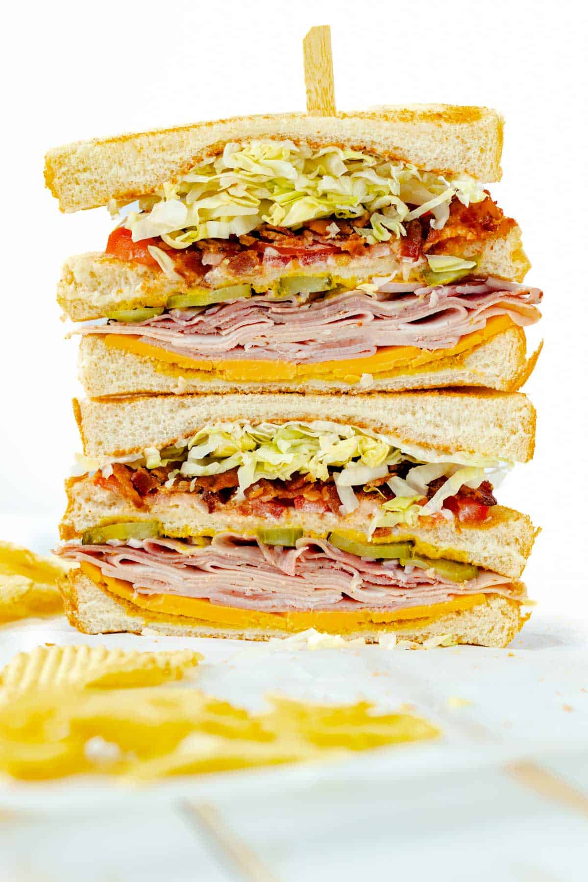 A classic Sports Dinners favorite: stacked ham, cheddar cheese, bacon, pickles, and shredded lettuce on white bread, held with a toothpick and served alongside crispy potato chips.