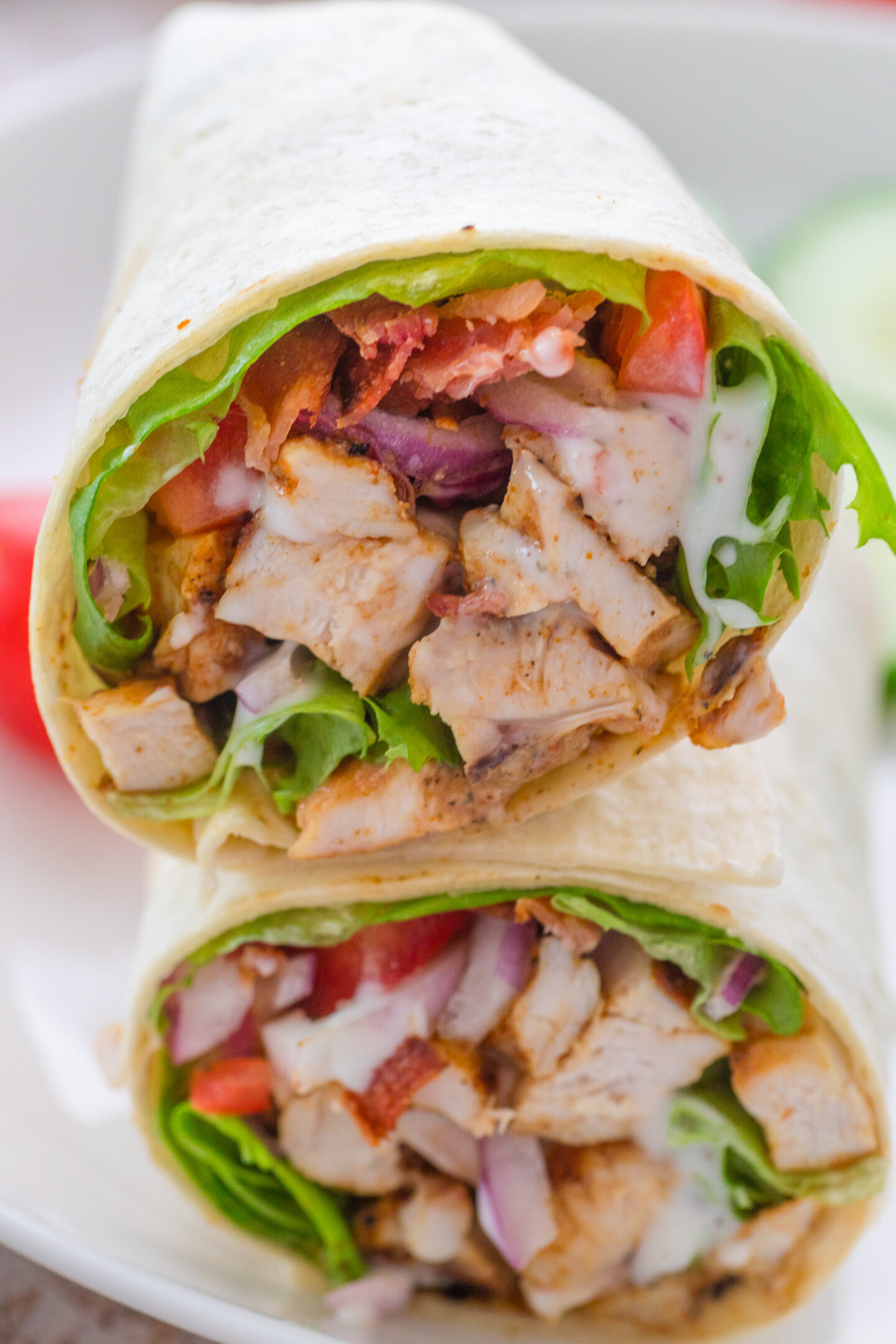 Two halves of a chicken wrap with lettuce, tomato, red onion, bacon, and a creamy sauce in a flour tortilla are served on a white plate—perfect for quick Sports Dinners.