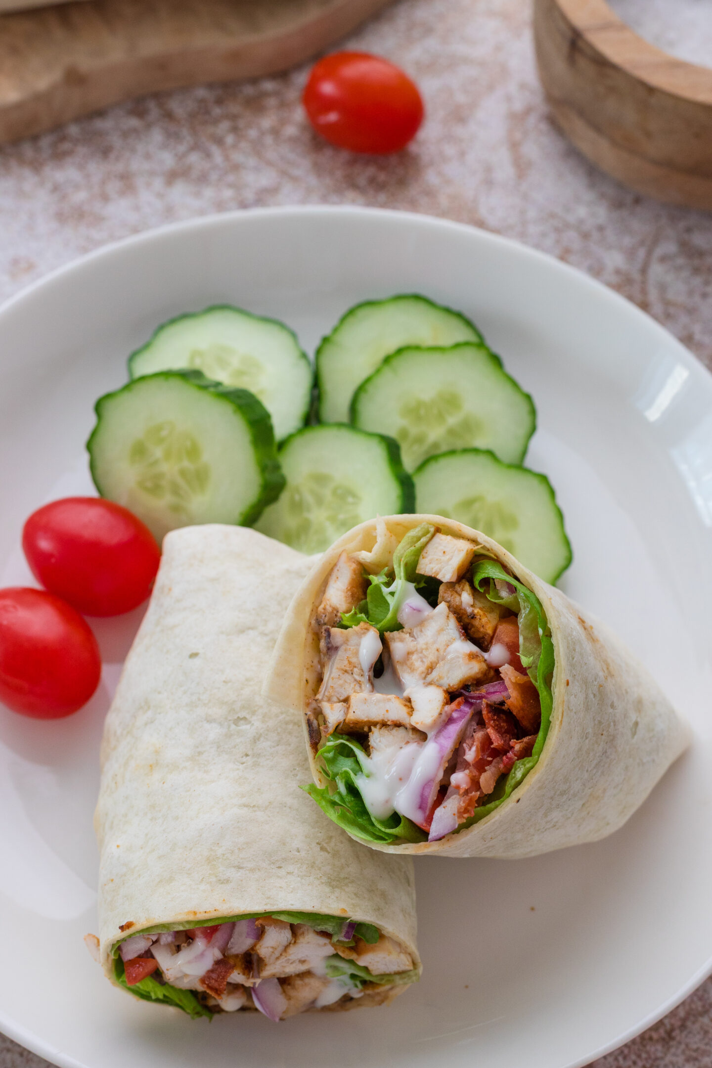 A plate with two halves of a chicken wrap, three slices of cucumber, and two cherry tomatoes—perfect for simple seaside dinner or beach meals.