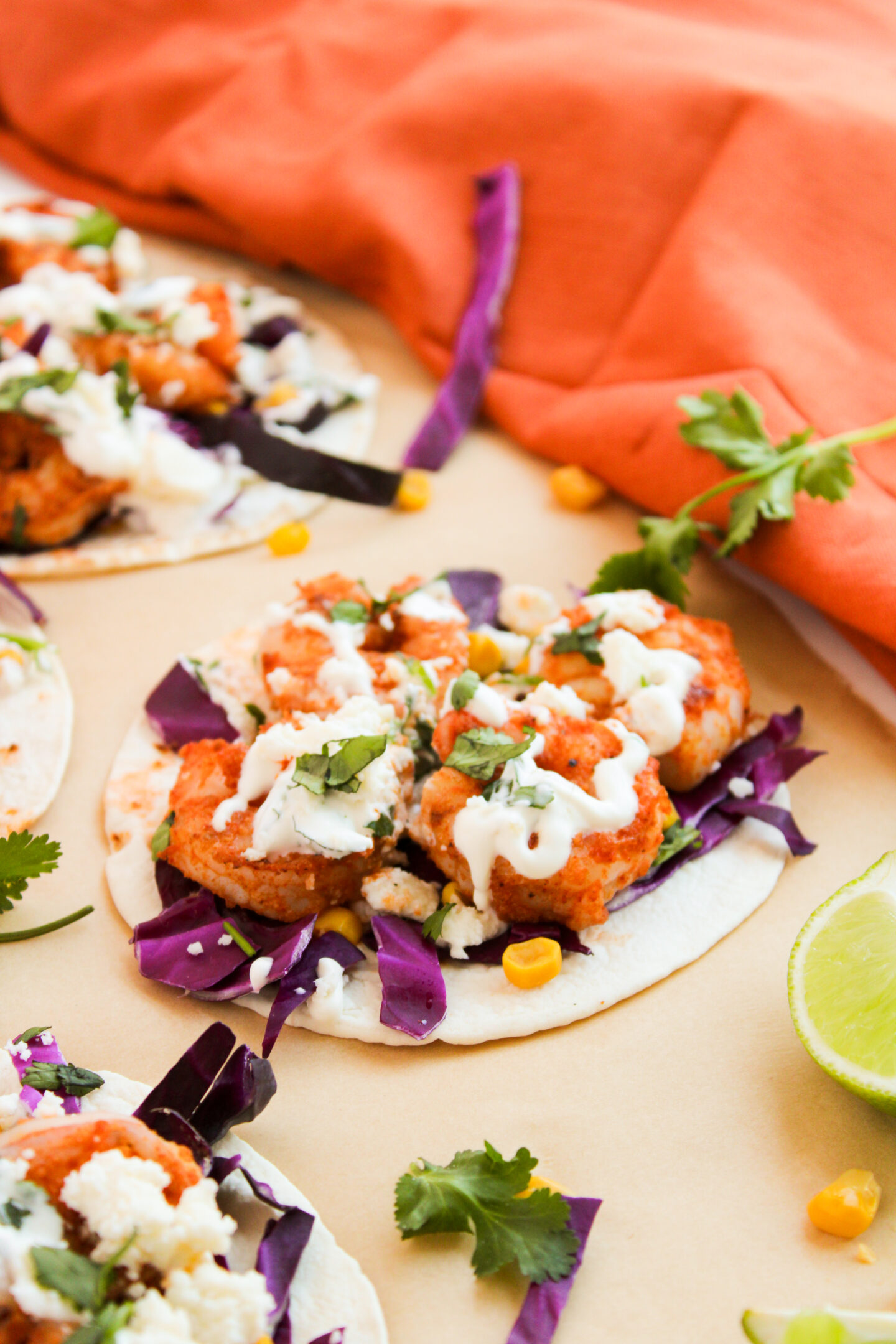 Three tacos topped with fried shrimp, purple cabbage, corn, cheese, chopped cilantro, and a drizzle of white sauce make perfect beach dinner ideas, served on a tan surface with a lime wedge and orange cloth nearby.