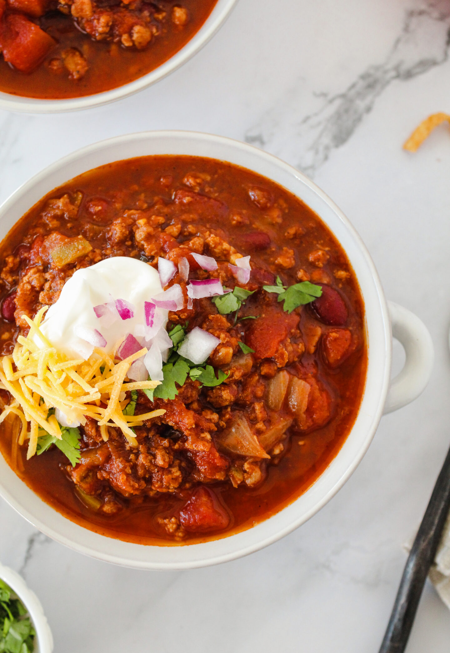 A bowl of chili topped with shredded cheese, sour cream, chopped red onions, and cilantro sits on a marble surface—perfect for cozy Sports Dinners.