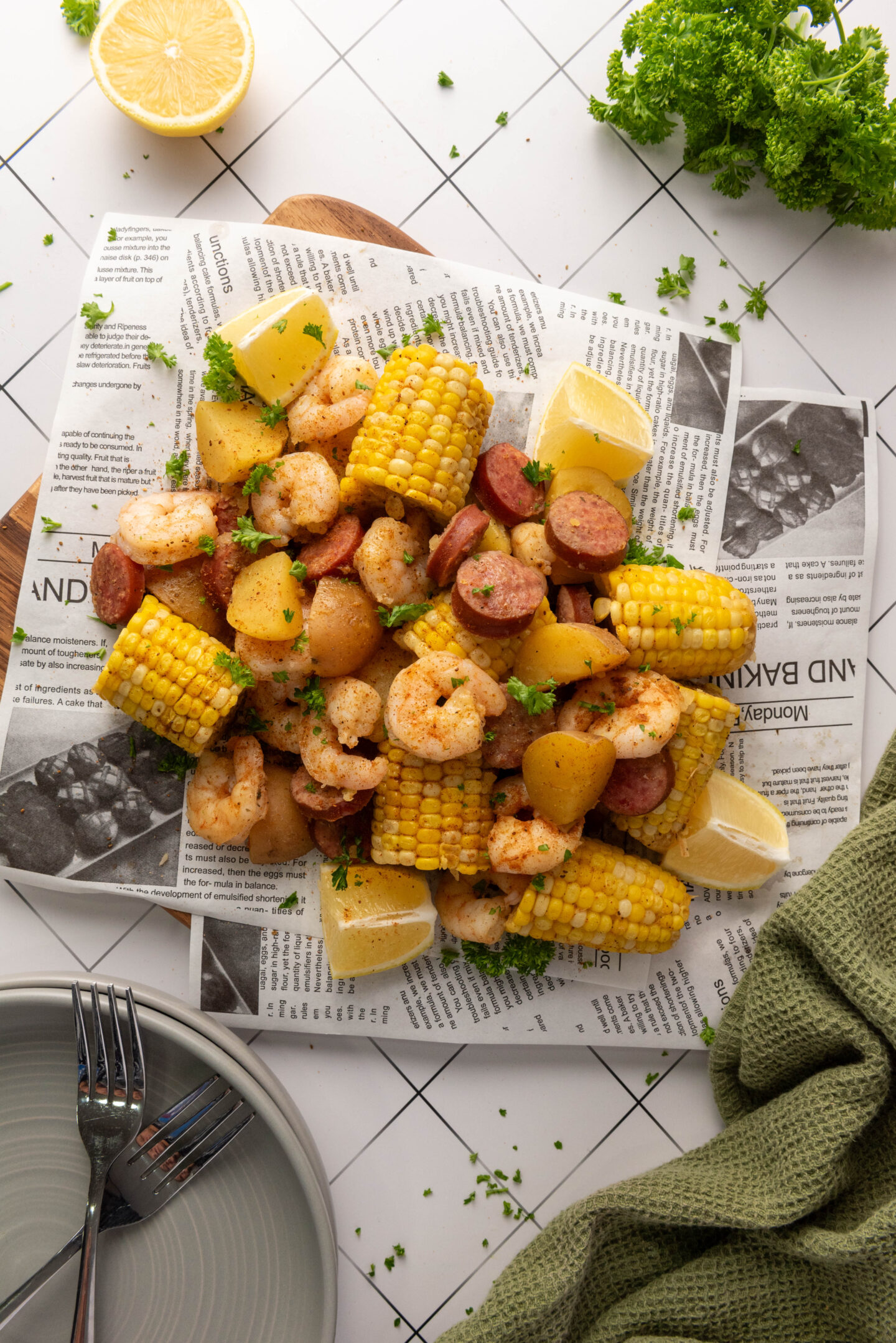A platter of shrimp, sausage, potatoes, and corn on the cob, garnished with lemon wedges and fresh herbs—an easy beach dinner served on newspaper atop white tiles, perfect for those seeking fun dinner ideas.