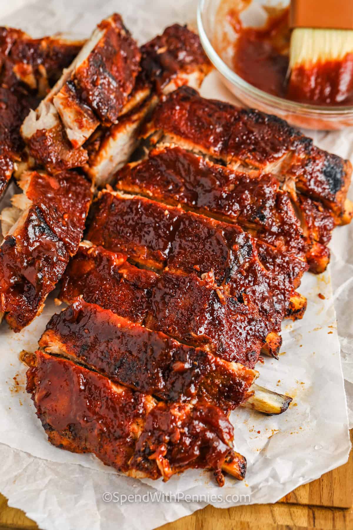 A rack of barbecued ribs sliced into pieces, coated in glossy barbecue sauce—perfect for a Father's Day Dinner—with a bowl of sauce and a basting brush in the background.