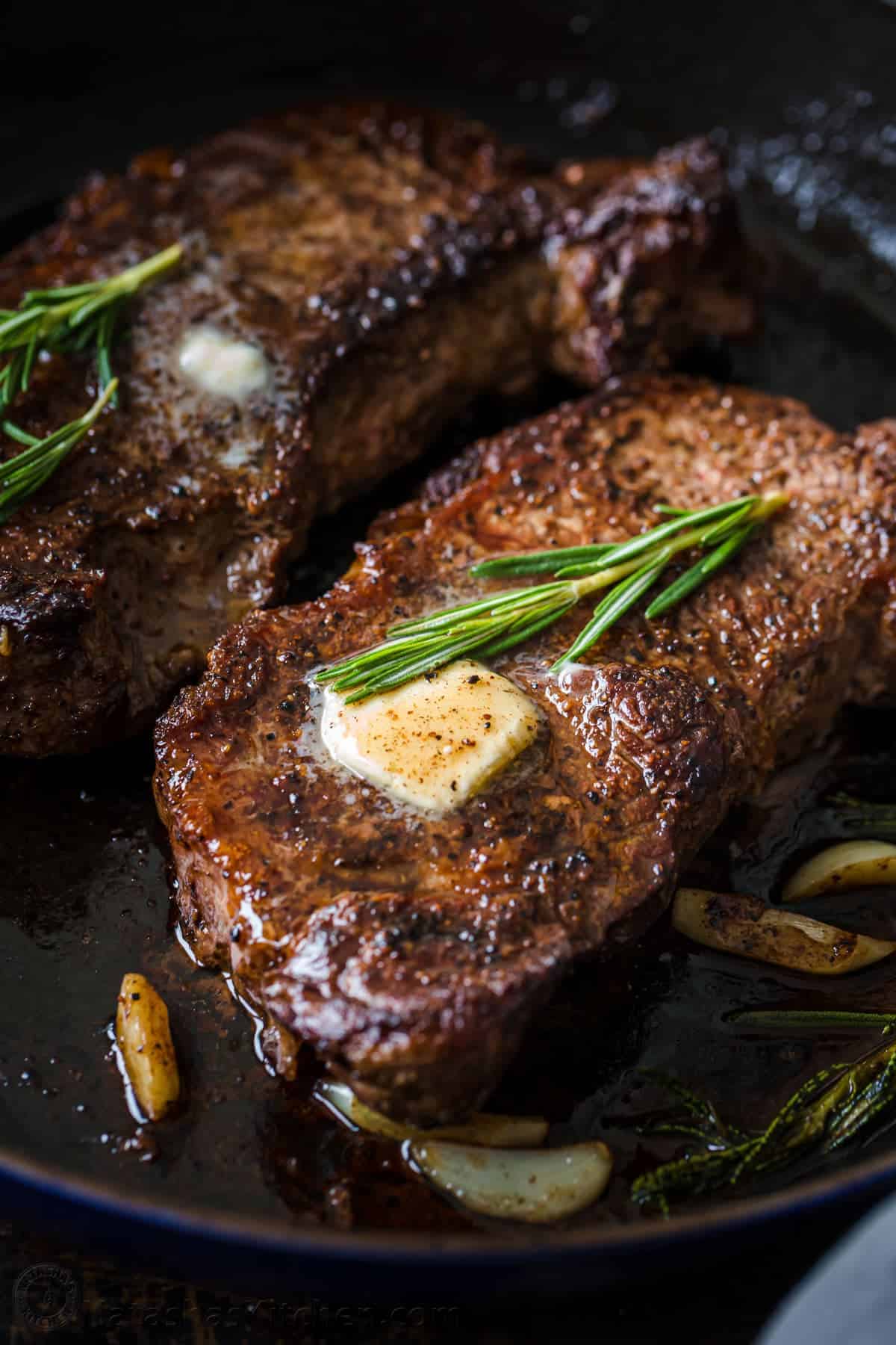 Two seared steaks in a pan, topped with melting butter and fresh rosemary, with garlic cloves visible around the steaks—a perfect Father’s Day dinner idea for celebrating dad.