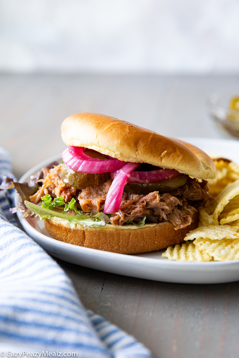 A pulled pork sandwich with lettuce, pickles, and pickled red onions on a bun, served with ridged potato chips on a white plate—a perfect beach dinner or addition to your list of summer dinner ideas.